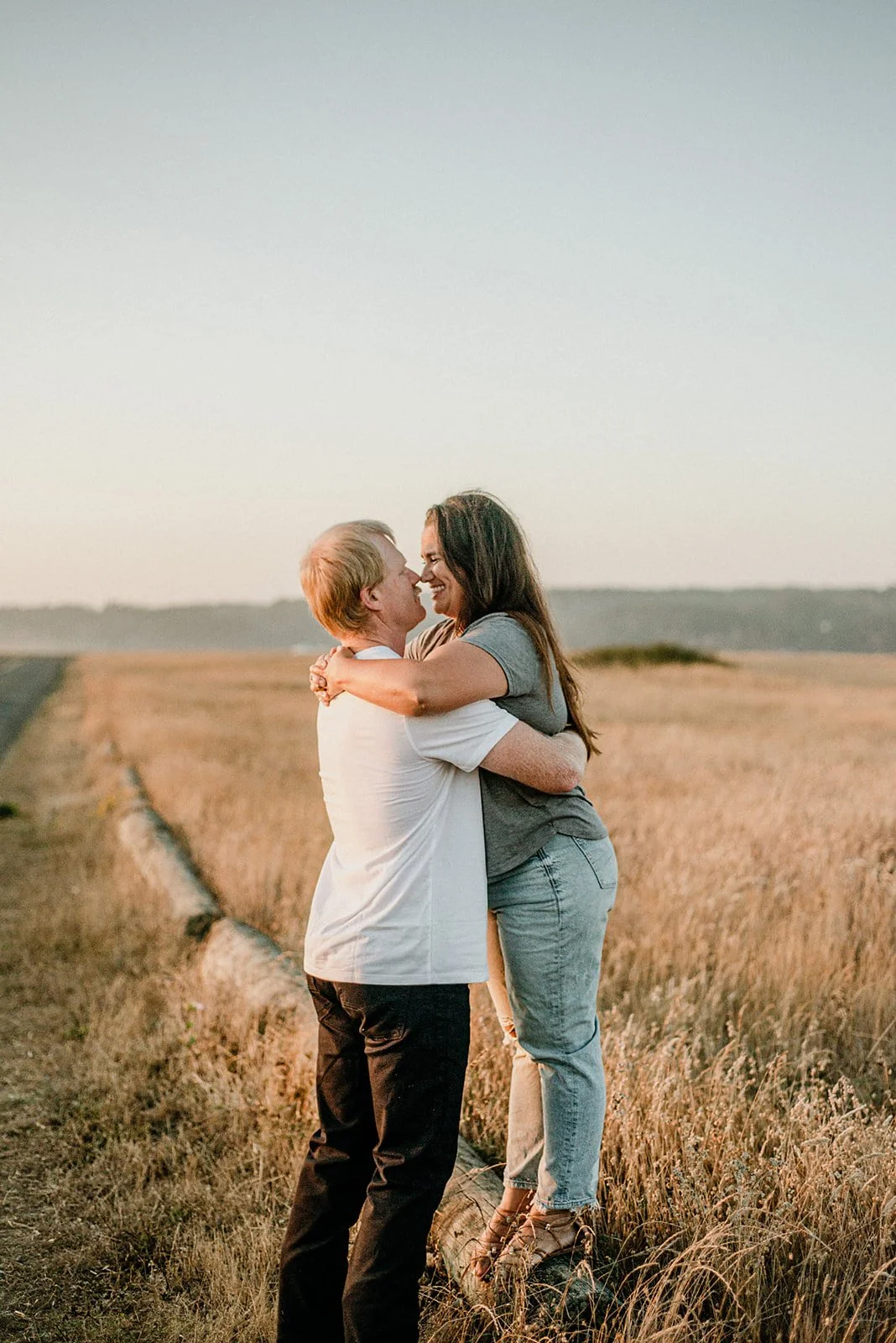 A couple hugging and smiling in a field during sunset, with open land in the background.