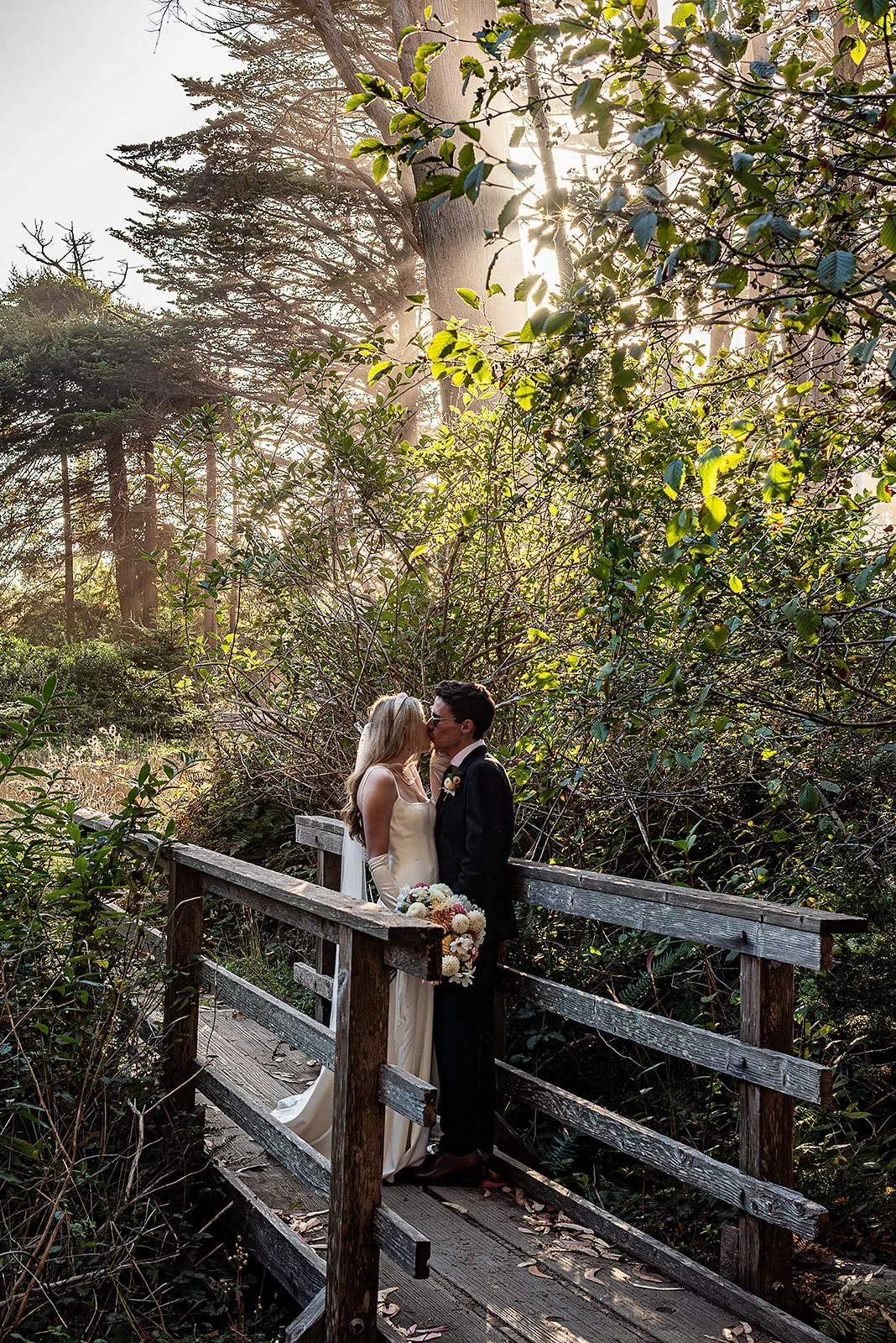 A bride and groom kissing on a wooden bridge surrounded by trees and greenery, with sunlight filtering through the leaves.