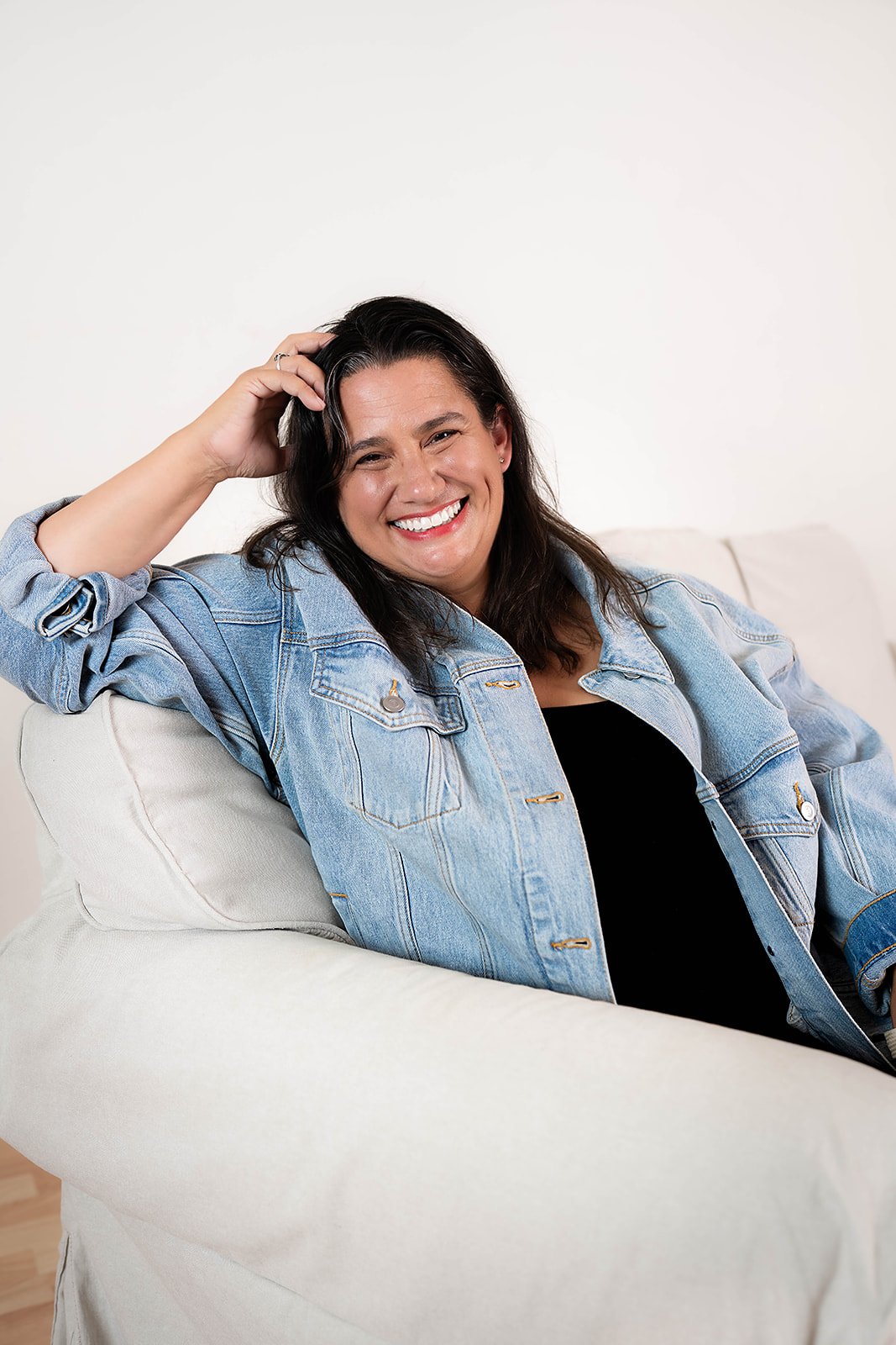 Smiling woman with dark hair, wearing denim jacket and black top, sitting on a light-colored couch against a white wall.
