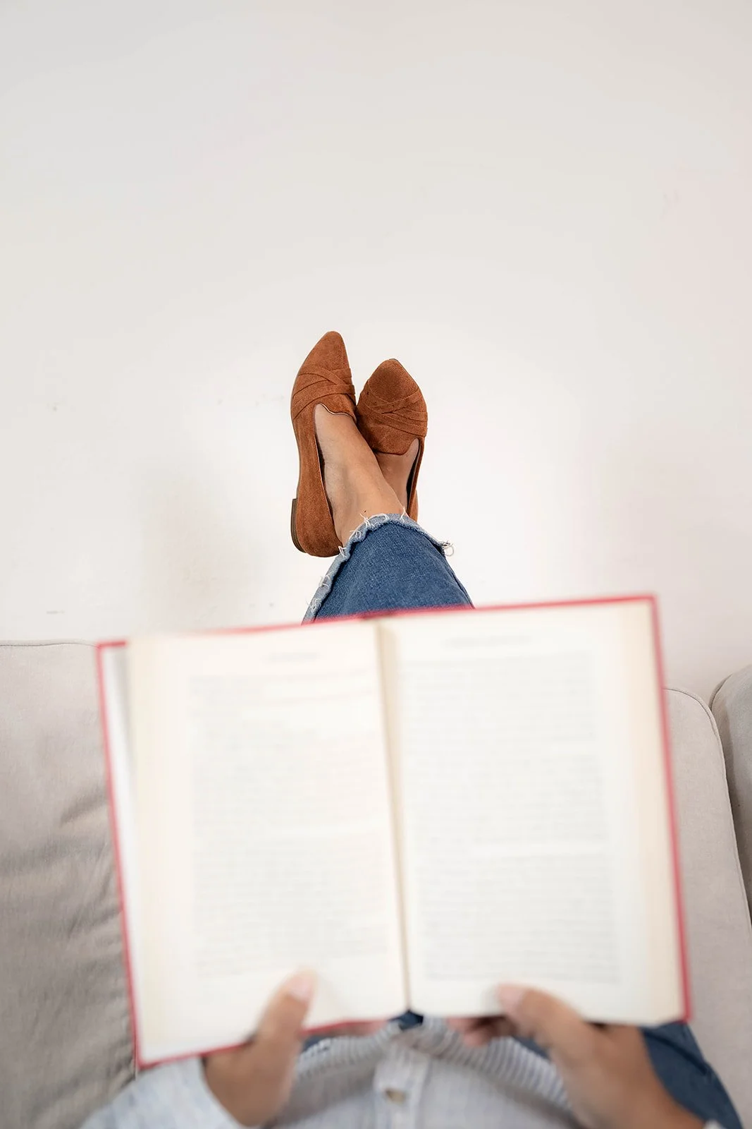 Person sitting on a sofa with legs crossed, wearing blue jeans and brown shoes, holding an open book, with a plain white background.