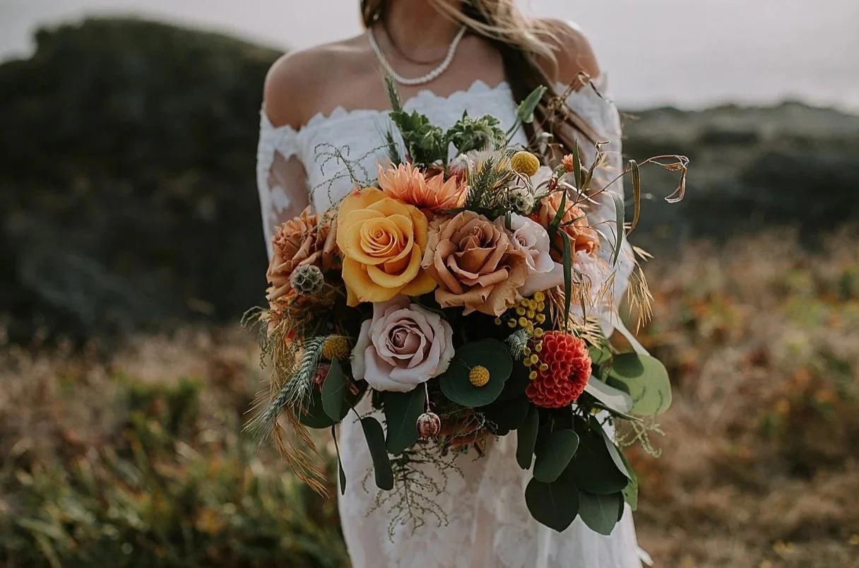 Woman in a white lace off-the-shoulder dress holding a large bouquet of flowers outdoors, with blurred natural landscape in the background.