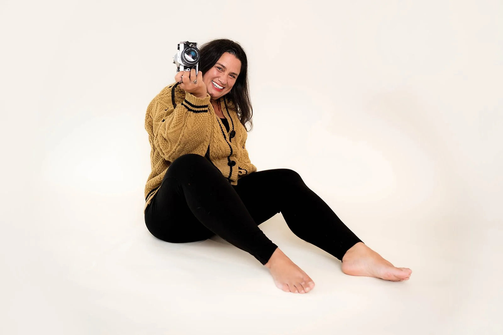 A woman sitting on the floor, barefoot, wearing a mustard-colored cardigan and black pants, smiling while holding a camera pointed towards the viewer.