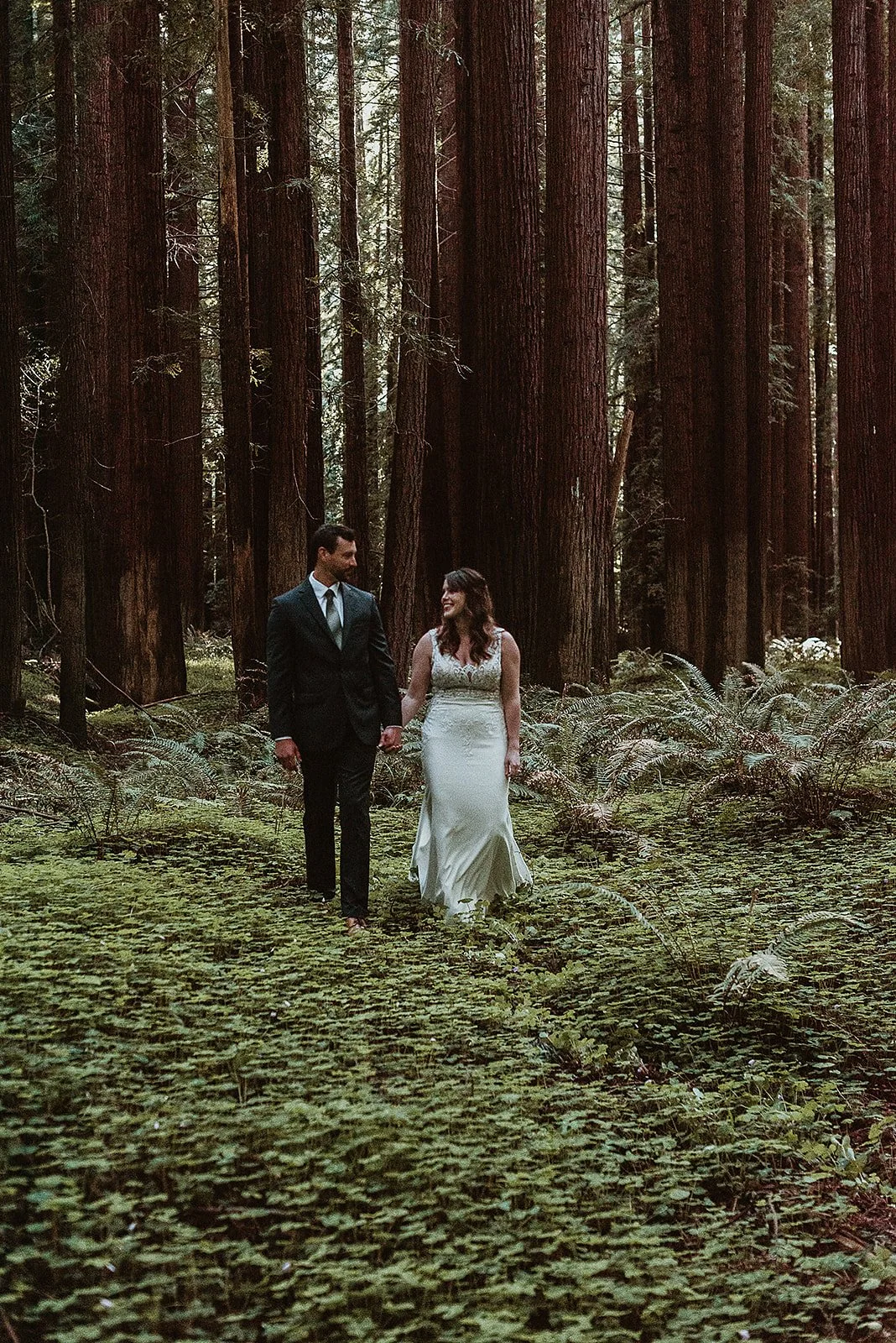 A bride and groom holding hands and walking through a forest with tall trees and green foliage.