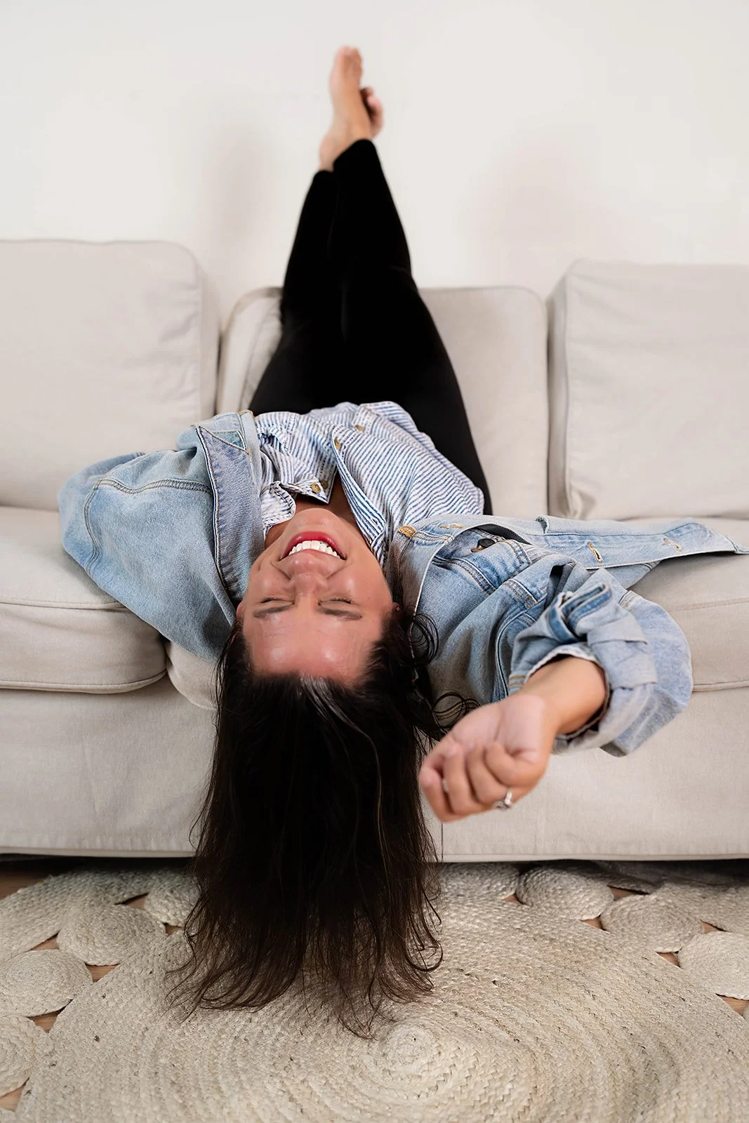 A woman lying upside down on a beige couch, smiling with her eyes closed, wearing a denim jacket and black pants, with a beige rug underneath.