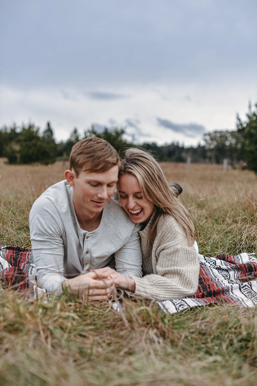 A couple lying on a checkered blanket in a grassy field, sharing a joyful moment together, with trees and cloudy sky in the background.