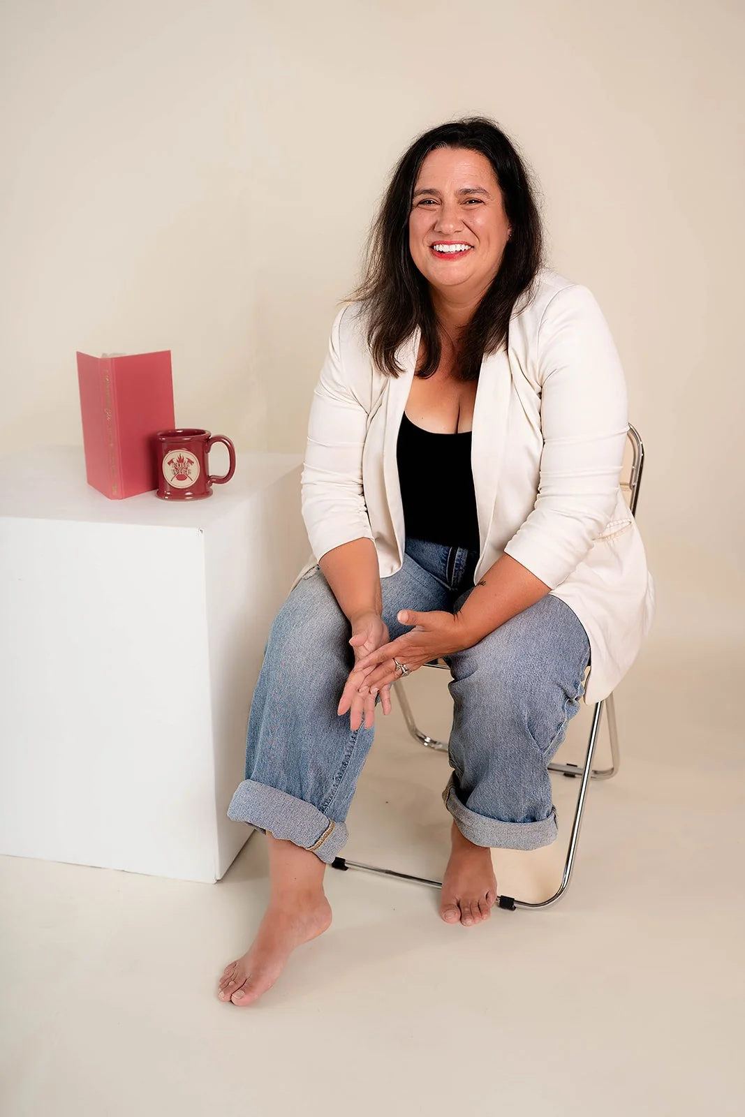 A smiling woman with dark hair, wearing a white blazer, black top, and cuffed jeans, sitting barefoot on a chair. Beside her, a white pedestal holds a pink book and a red mug with a logo.