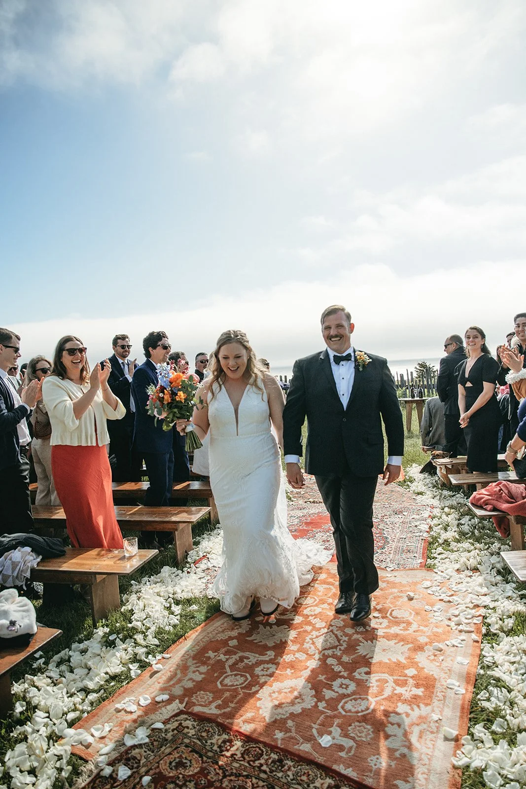 Bride and groom walking down the aisle outdoors after their wedding ceremony, surrounded by smiling guests clapping and taking photos, on a bright day with a blue sky.