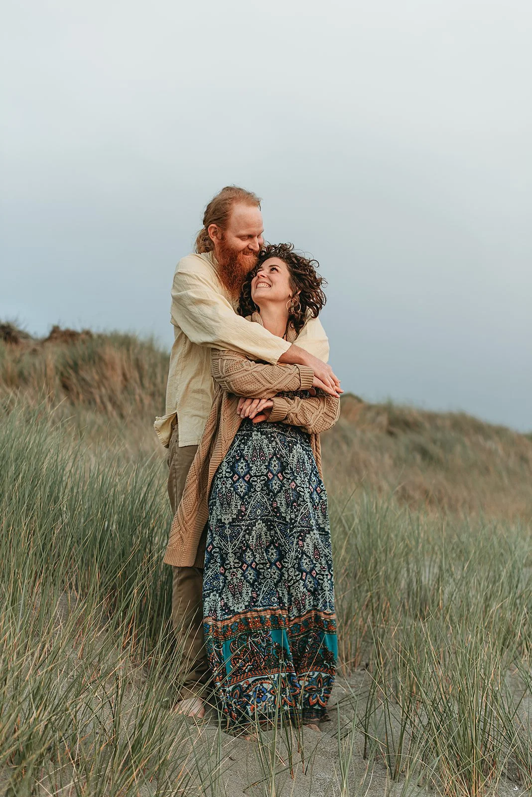 A couple embracing on a grassy beach, smiling and looking into each other's eyes, with dunes and a cloudy sky in the background.