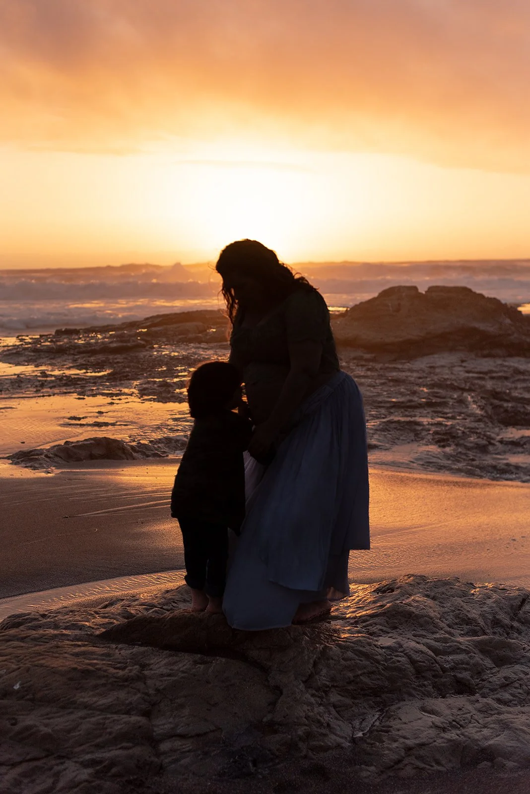 A silhouette of a woman and a small child standing on the beach at sunset, facing each other with the ocean and a vibrant orange sky in the background.