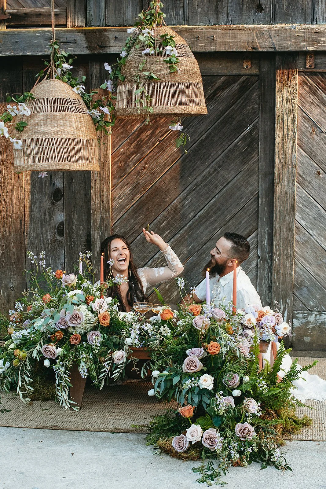 A couple sitting at a table decorated with an abundance of pastel-colored roses, greenery, and flowers, celebrating with lit candles in a rustic outdoor setting with wooden boards and hanging wicker lamps.