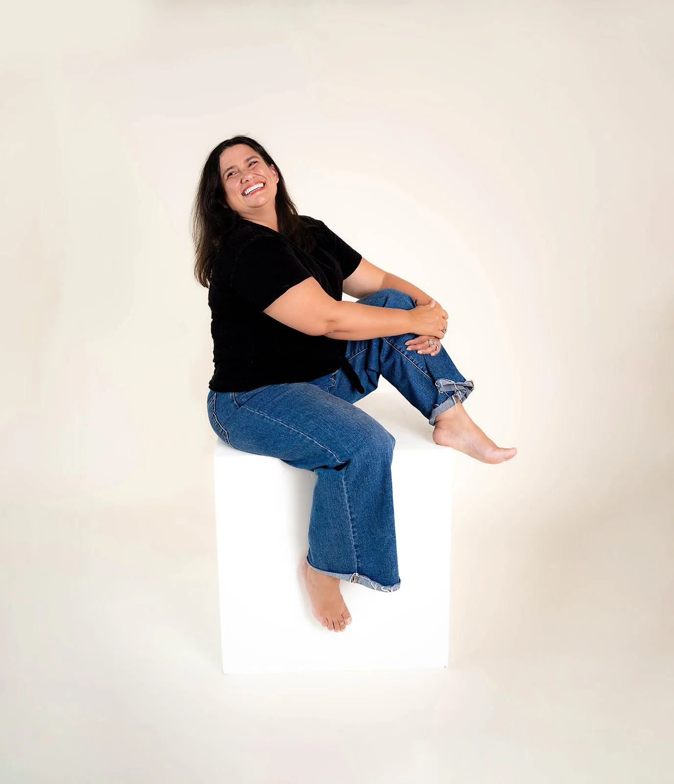 A woman with dark hair, wearing a black t-shirt and blue jeans, sitting barefoot on a white cube with one leg crossed over the other, laughing against a plain light-colored background.