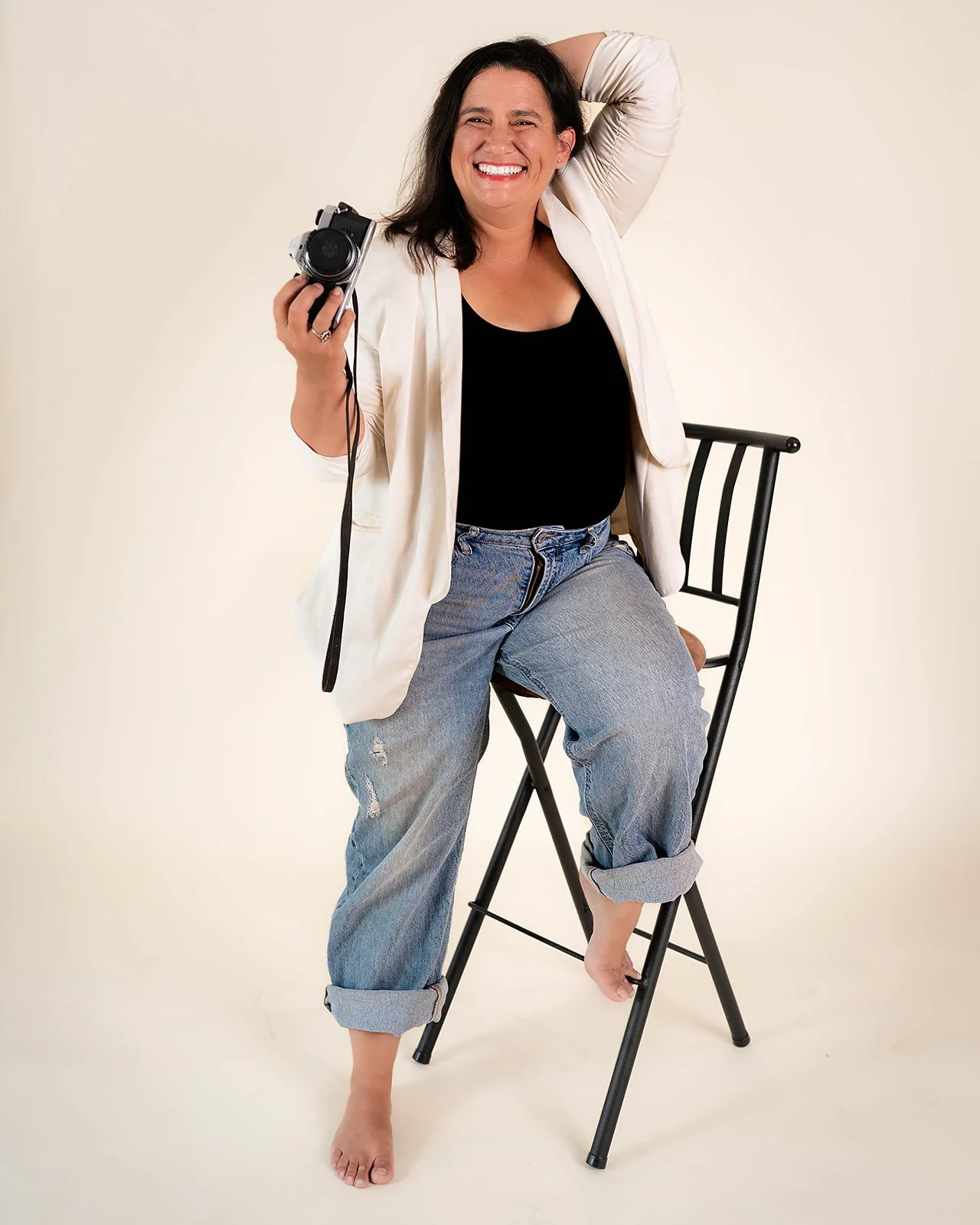 A woman smiling in casual attire sitting on a chair, holding a camera in her hand, with one foot on the chair's rung and the other foot barefoot on the floor.