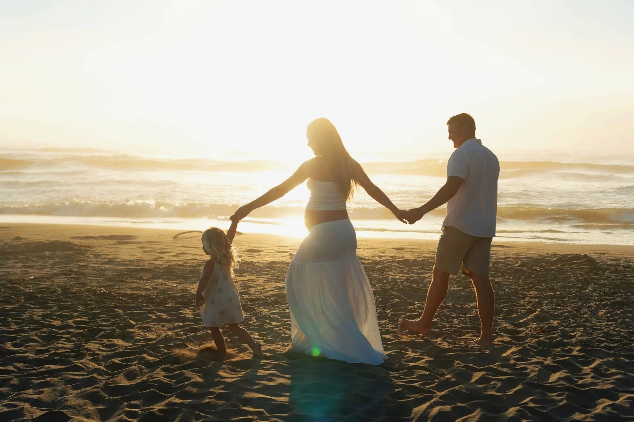 A family walking hand in hand on the beach during sunset, with the mother, father, and young daughter silhouetted against the bright horizon.