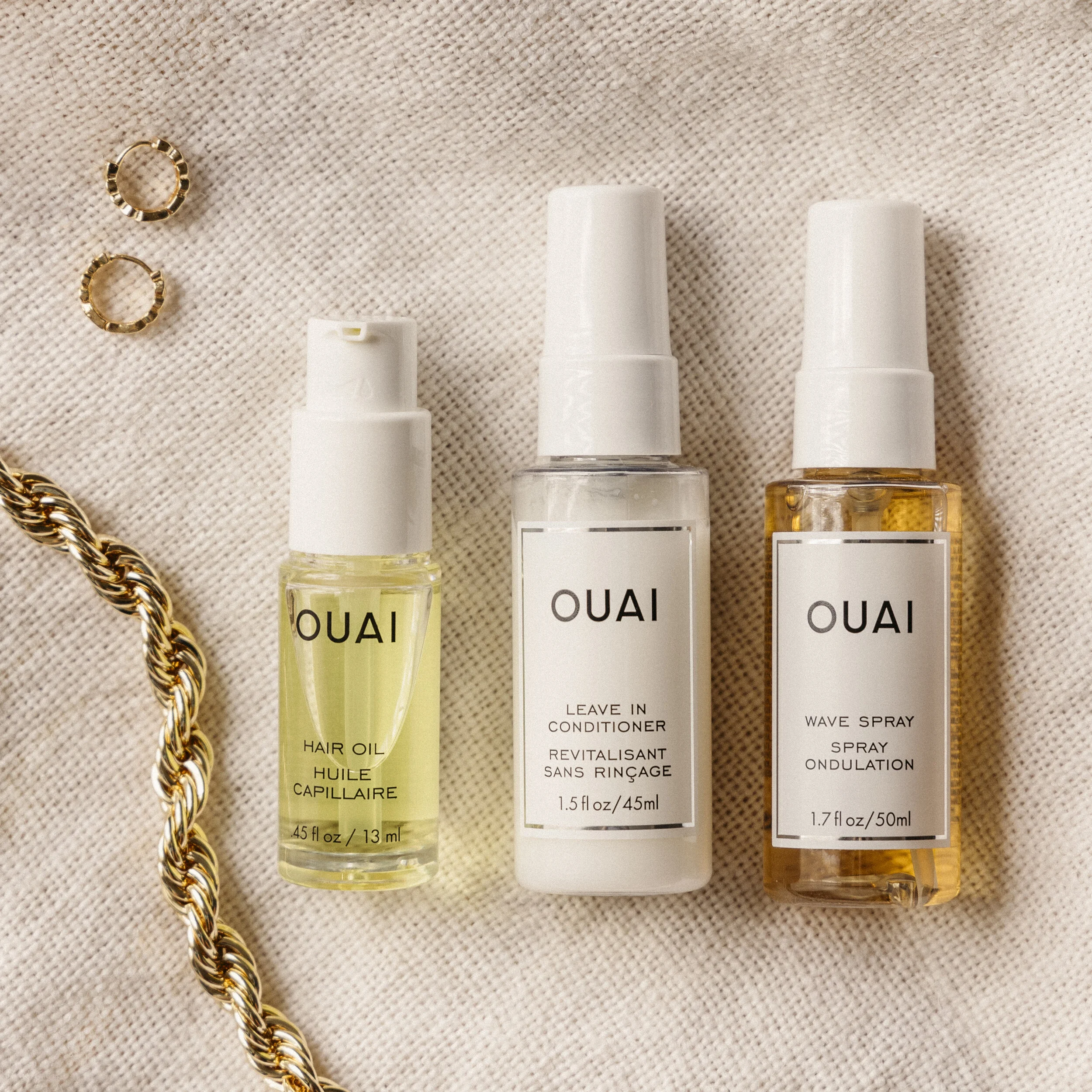 Three bottles of OUAI hair products: hair oil, leave-in conditioner, and wave spray, along with a gold chain and two small gold rings, all on a beige fabric background.