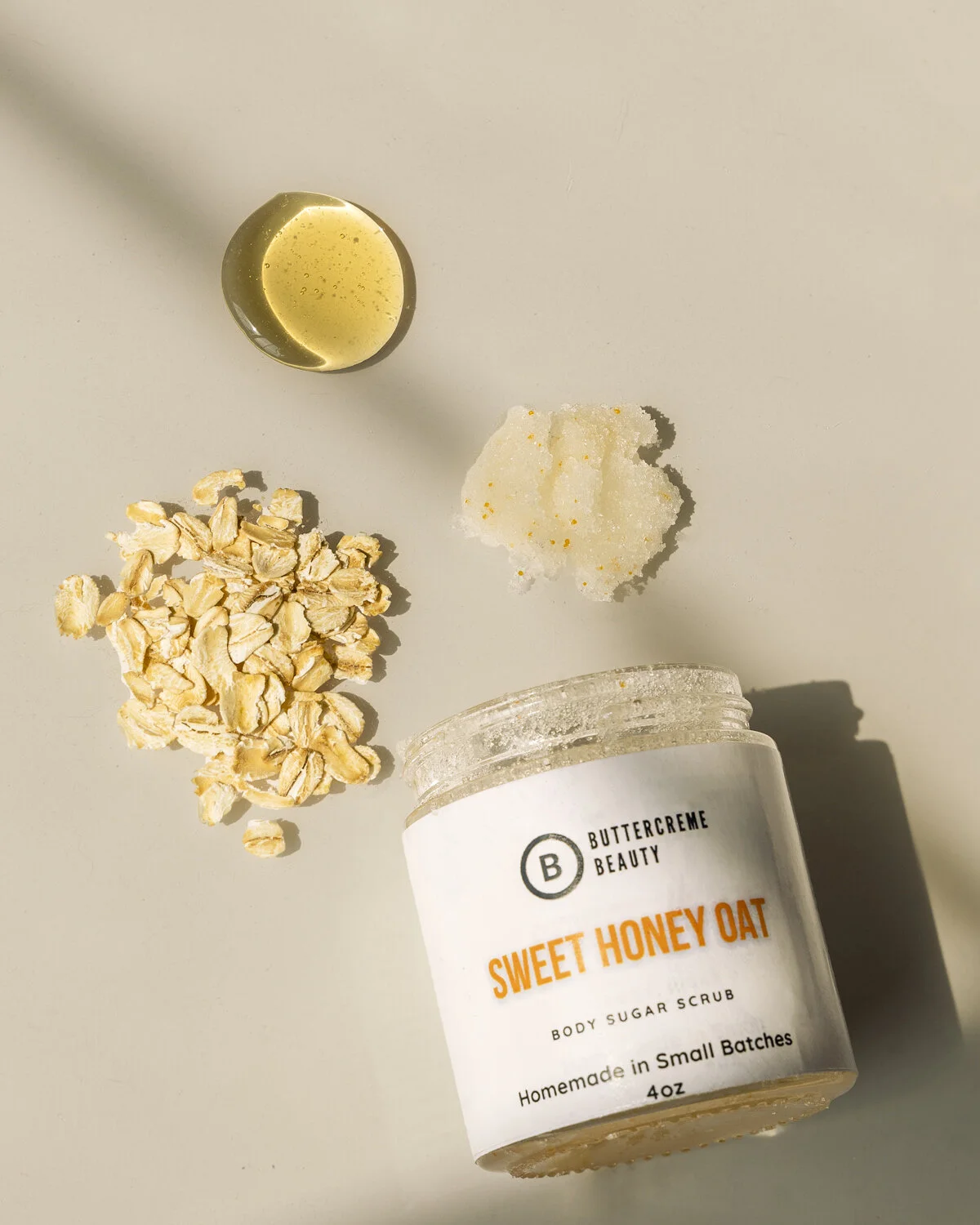Containers and ingredients for a body sugar scrub. A jar labeled 'Sweet Honey Oat' with oatmeal, honey, and a white scrub next to it. The jar is open, showing the scrub inside.