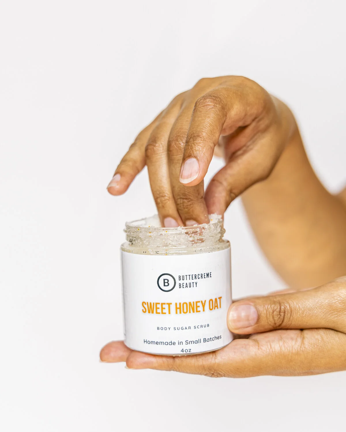 A person’s hand dipping fingers into a jar of Buttercreme Beauty Sweet Honey Oat body sugar scrub.