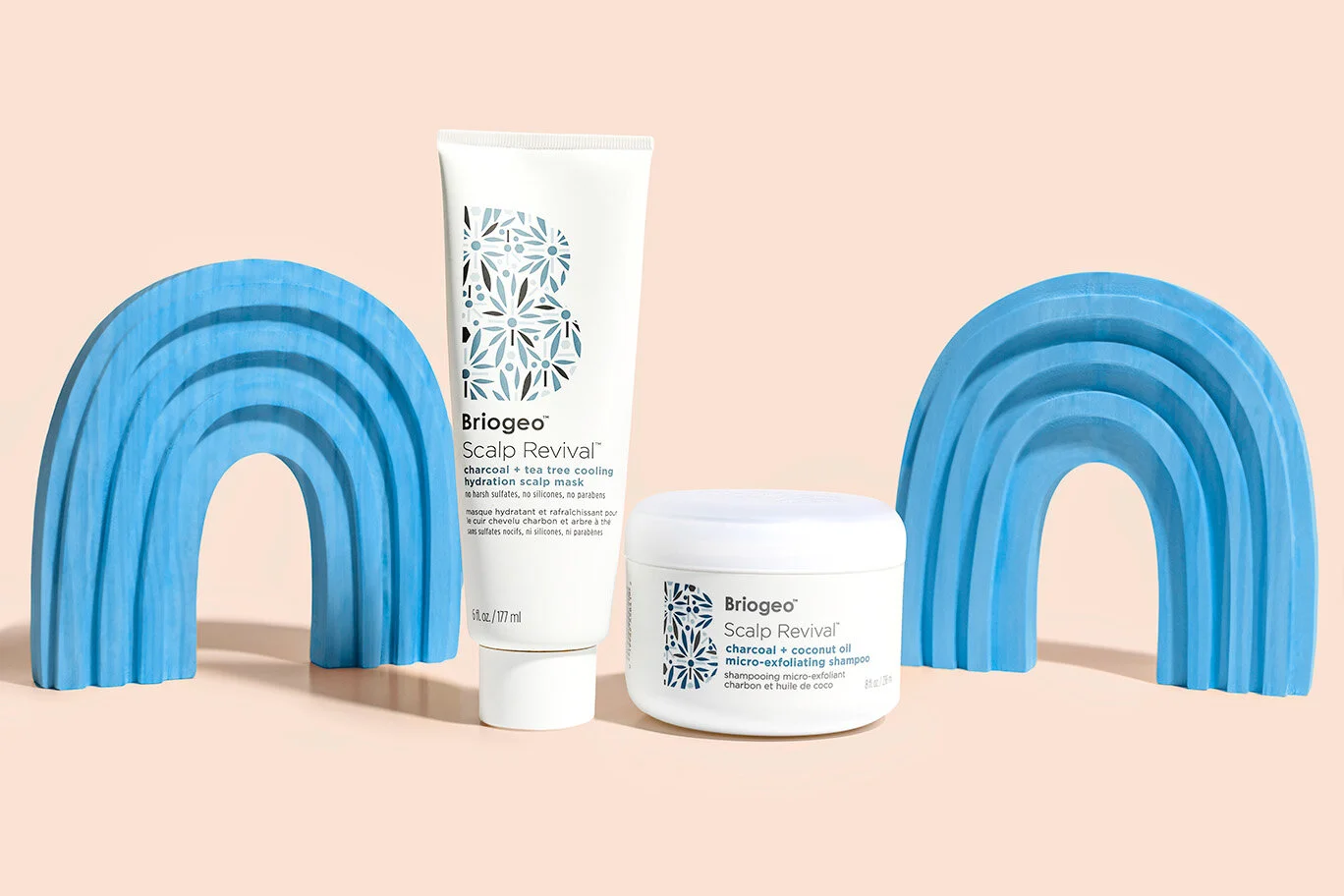Briogeo scalp revival products including a charcoal + tea tree cooling hydration scalp mask and a charcoal + coconut oil micro-exfoliating shampoo between two blue rainbow-shaped objects on a light pink background.