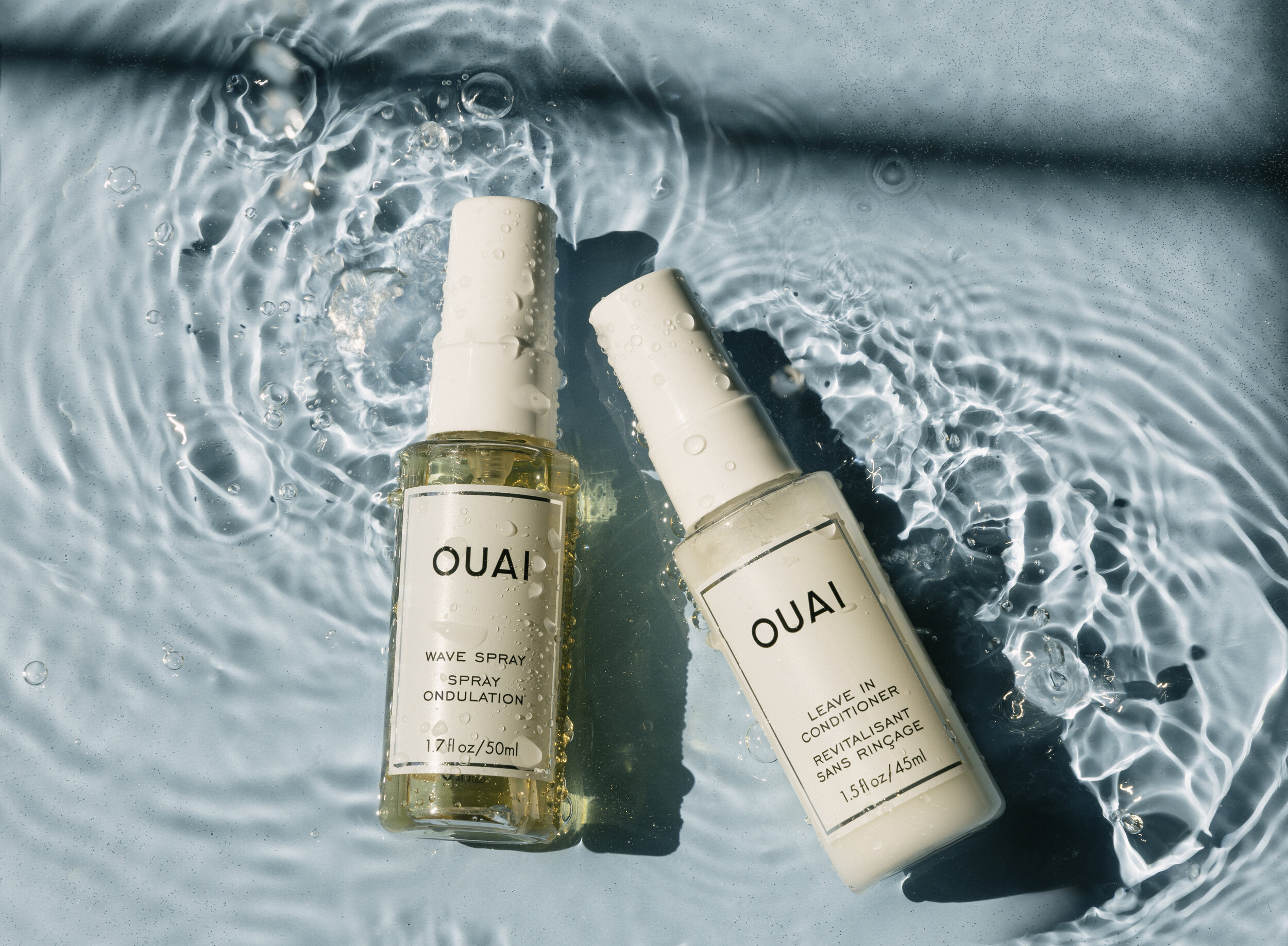Two bottles of OUAI hair products, one wave spray and one leave-in conditioner, floating on water with ripples and droplets.