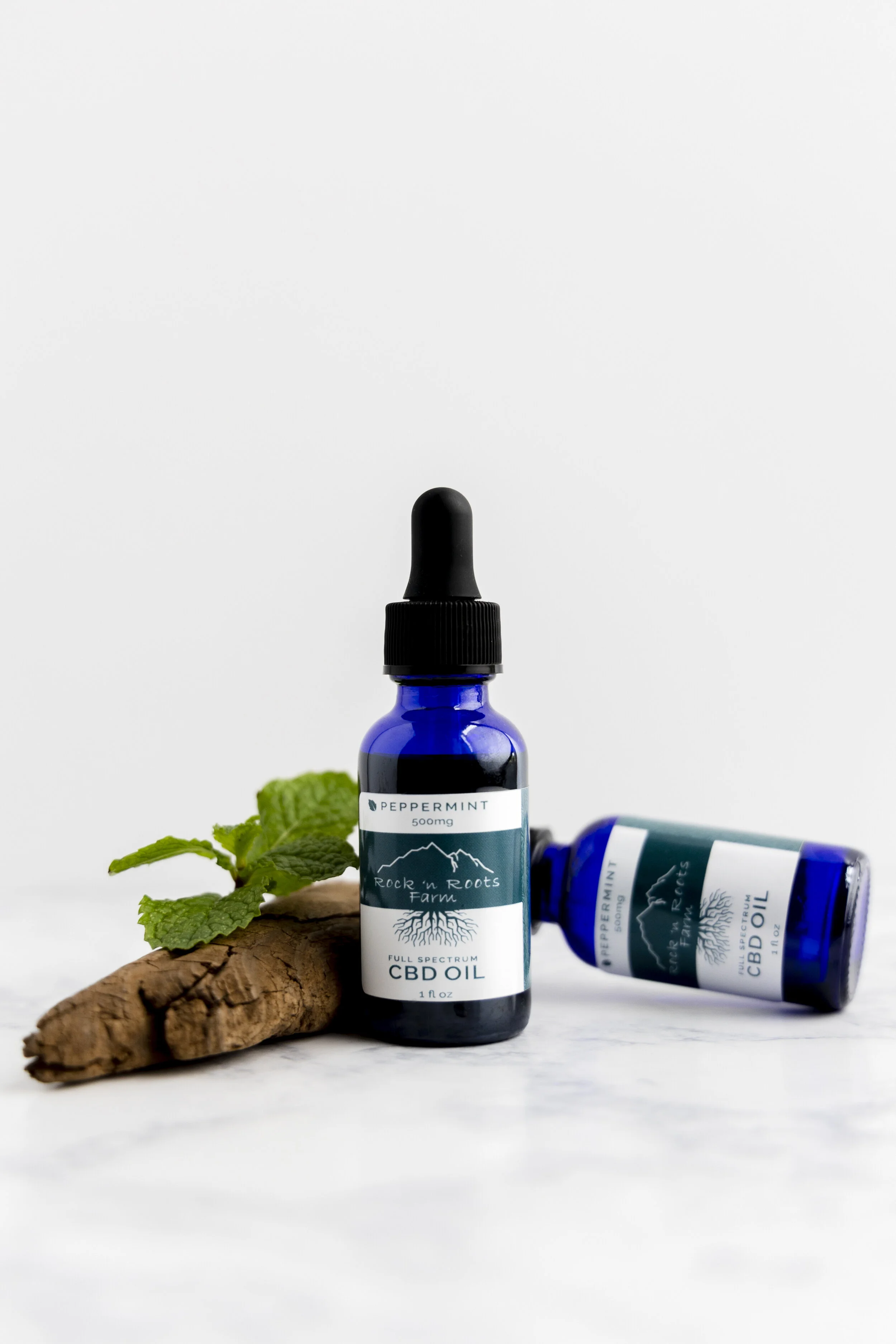 Two blue bottles of CBD oil with one upright and the other lying down, a piece of ginger root, and fresh mint leaves on a white surface.