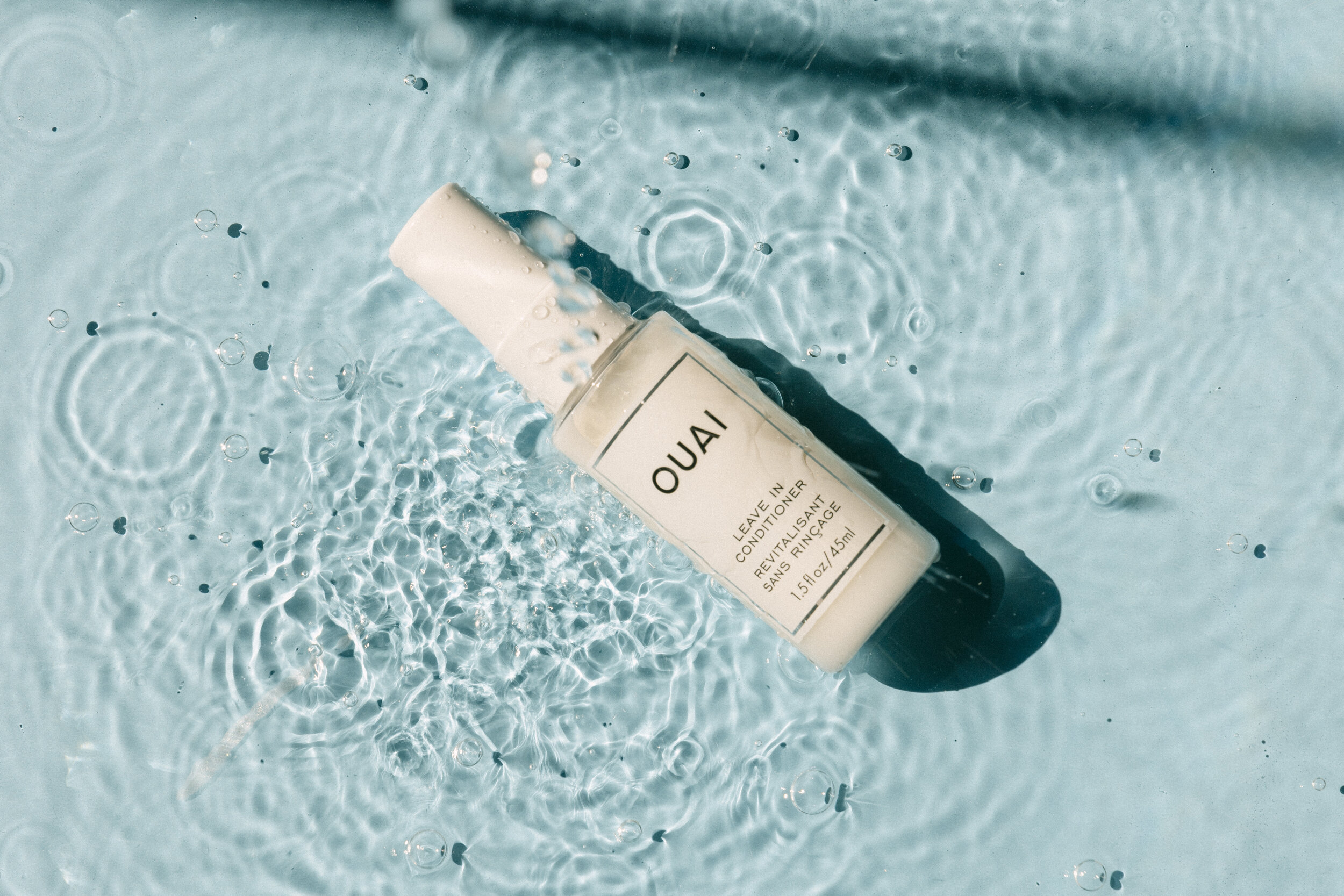 A white bottle of Ouai leave-in conditioner floating in light blue water with bubbles and ripples.