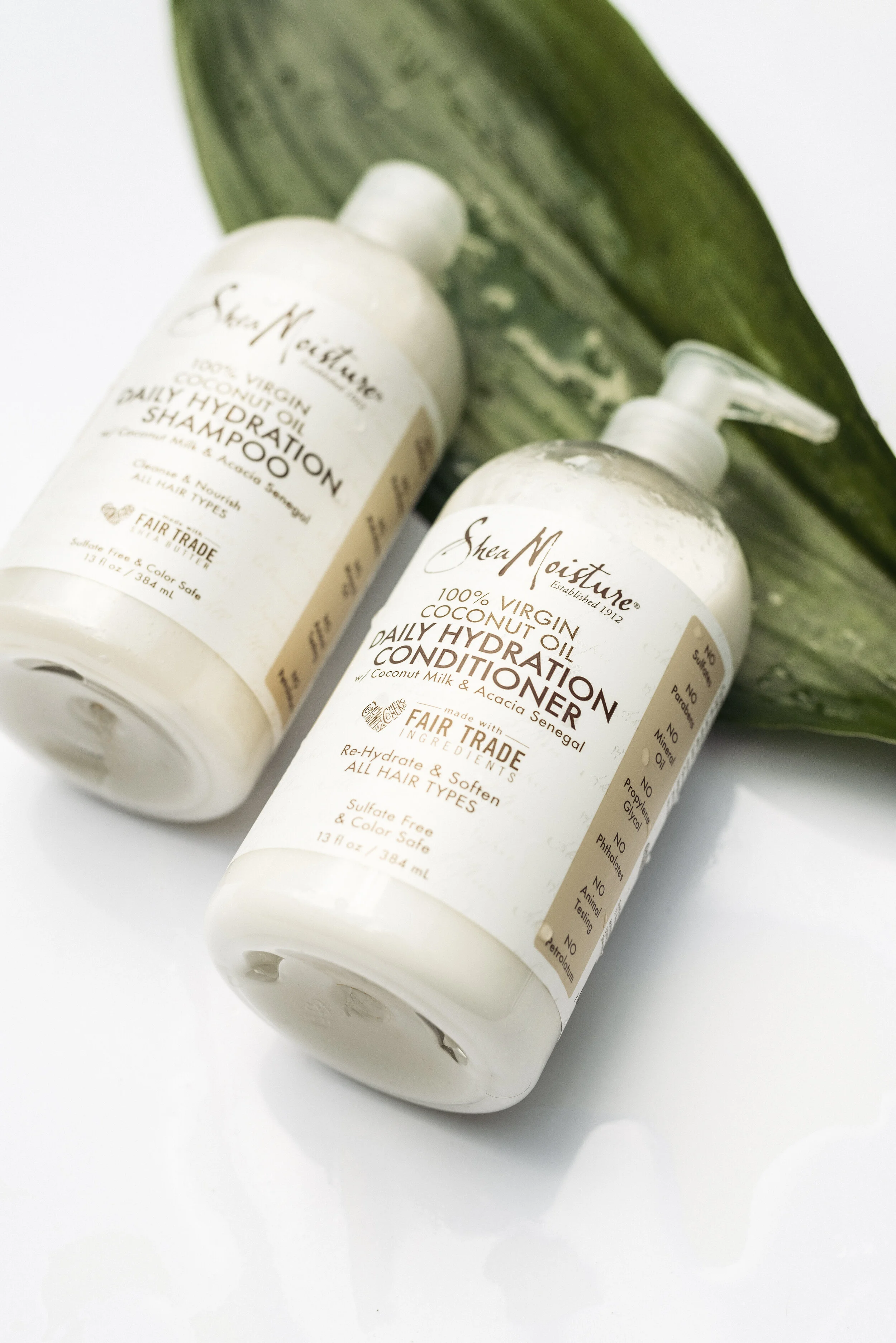 Two bottles of Shea Moisture daily hydration conditioner and shampoo with green leaves in the background.