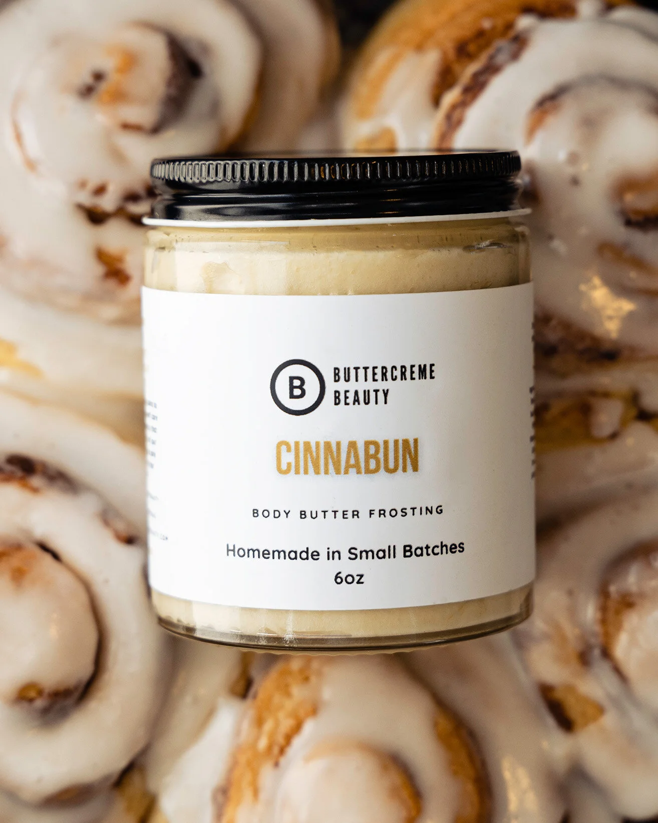 A jar of Buttercreme Beauty Cinnabon body butter frosting in front of sliced cinnamon rolls with icing.