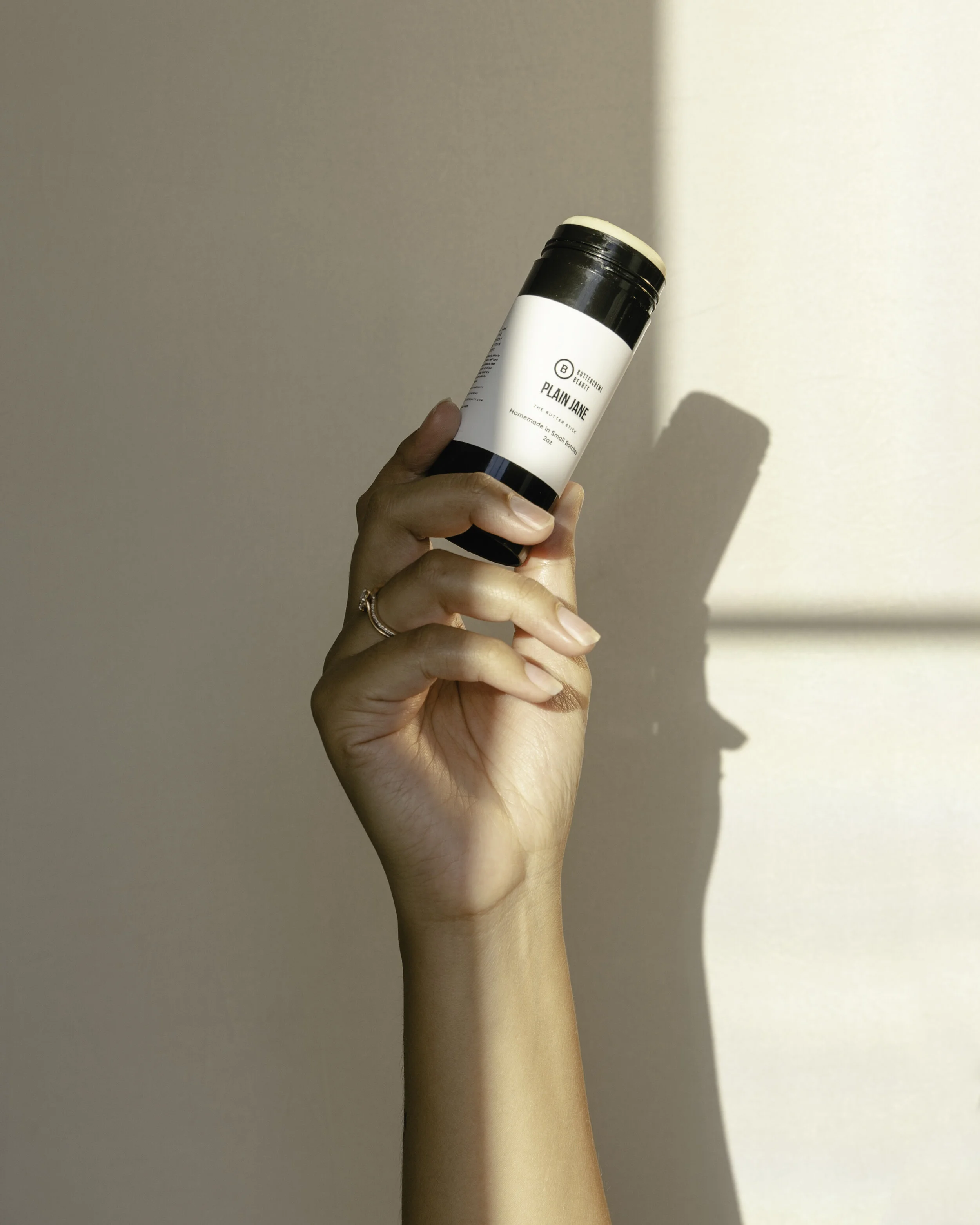 A hand holding a black and white container labeled 'Plain Jane' against a plain wall with sunlight casting a shadow.