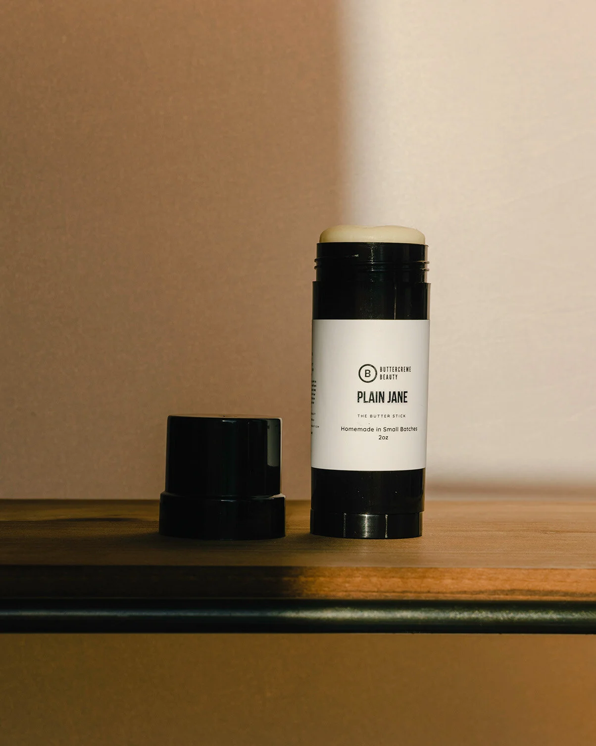 A black cylindrical container labeled 'Plain Jane' from Buttercreme Beauty, next to a smaller black lid, both placed on a wooden surface against a plain background.
