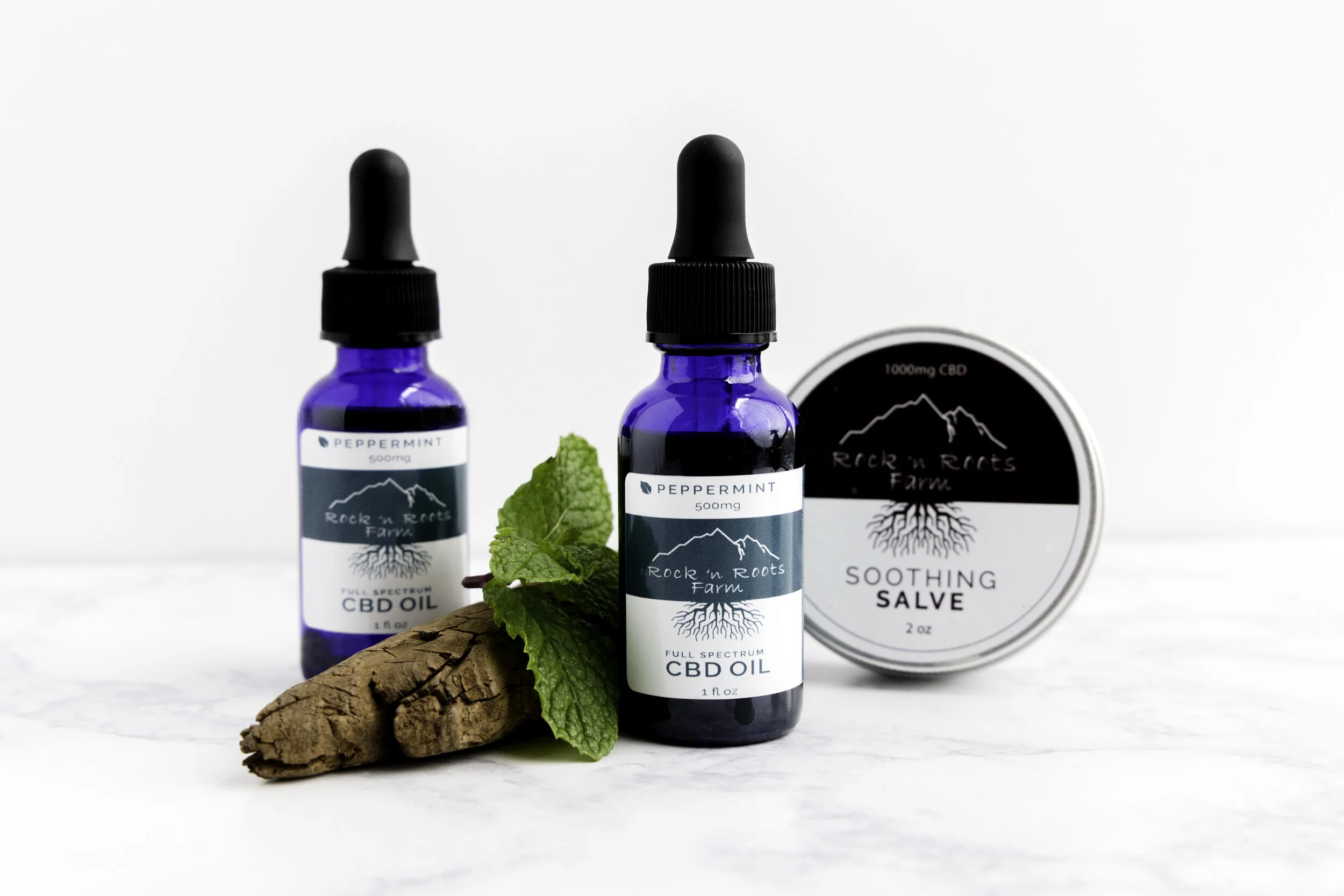 Two blue bottles of CBD oil with dropper caps, a sprig of fresh mint, a piece of ginger root, and a round container of soothing salve on a white surface.