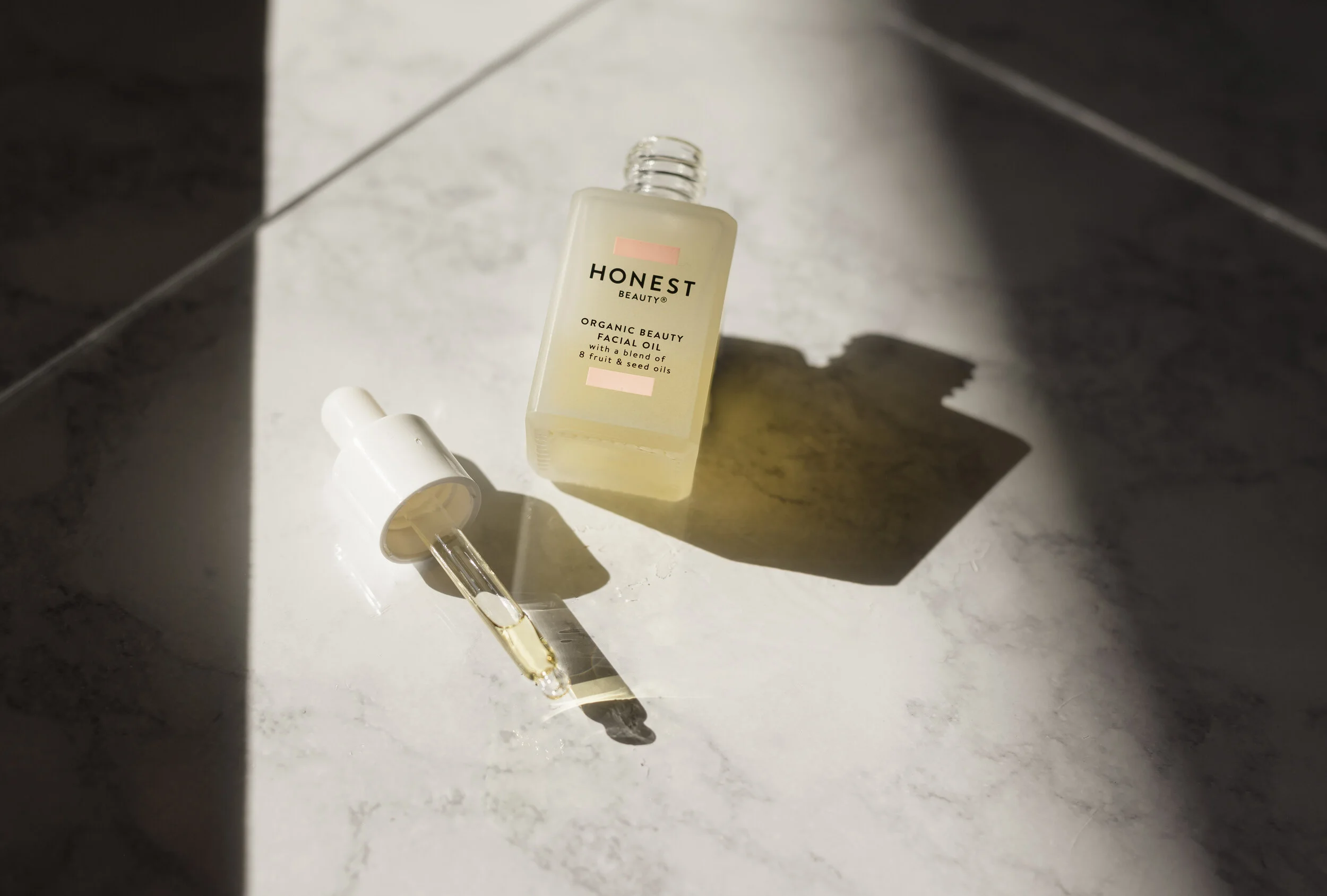 A bottle of Honest Beauty organic facial oil with a dropper cap, and a dropper containing some oil, placed on a marble surface in natural sunlight.