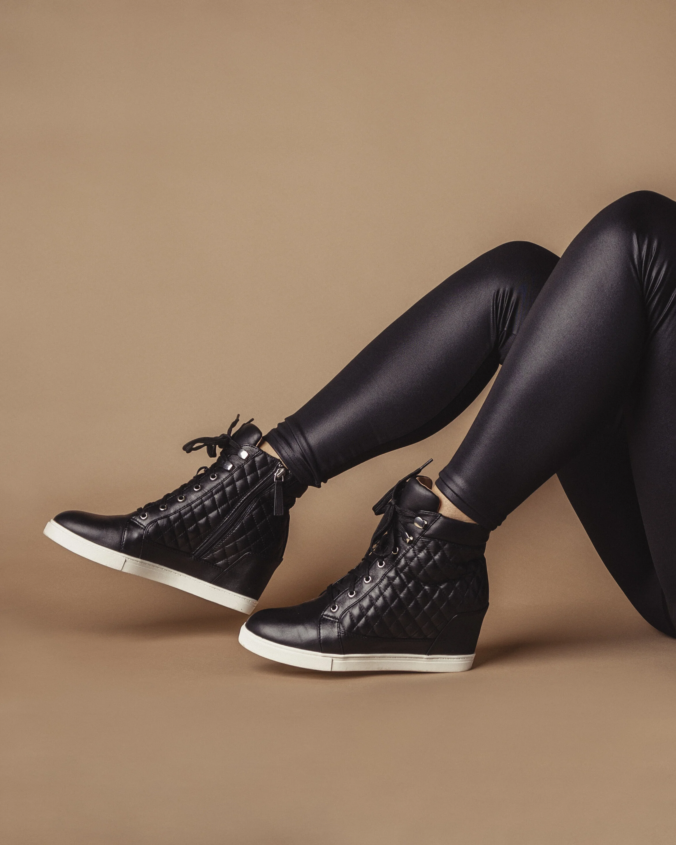 Person wearing black quilted wedge sneakers and shiny black leggings, lying against a beige background.