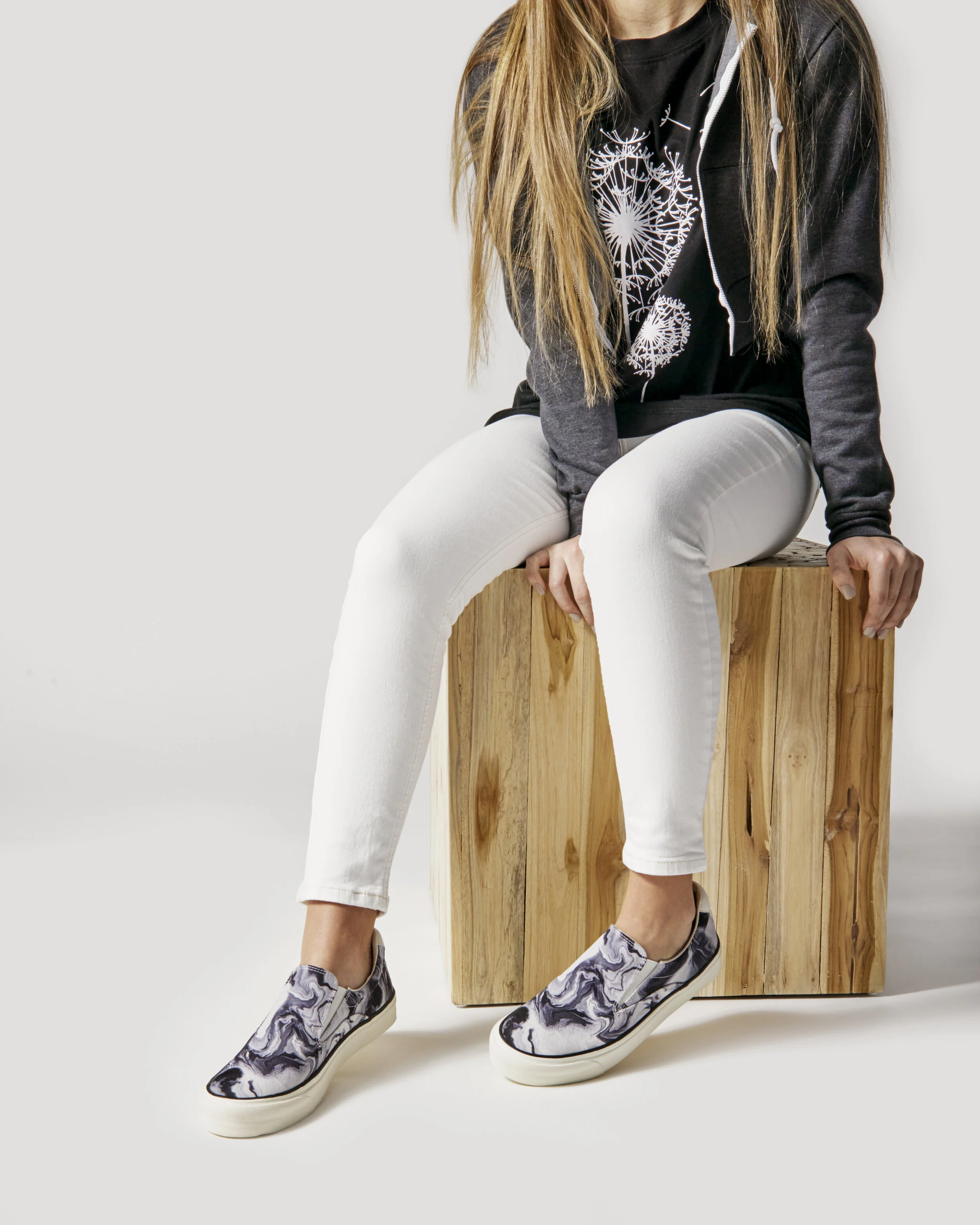 Person sitting on a wooden block wearing a black jacket, white pants, and marble-patterned slip-on sneakers.