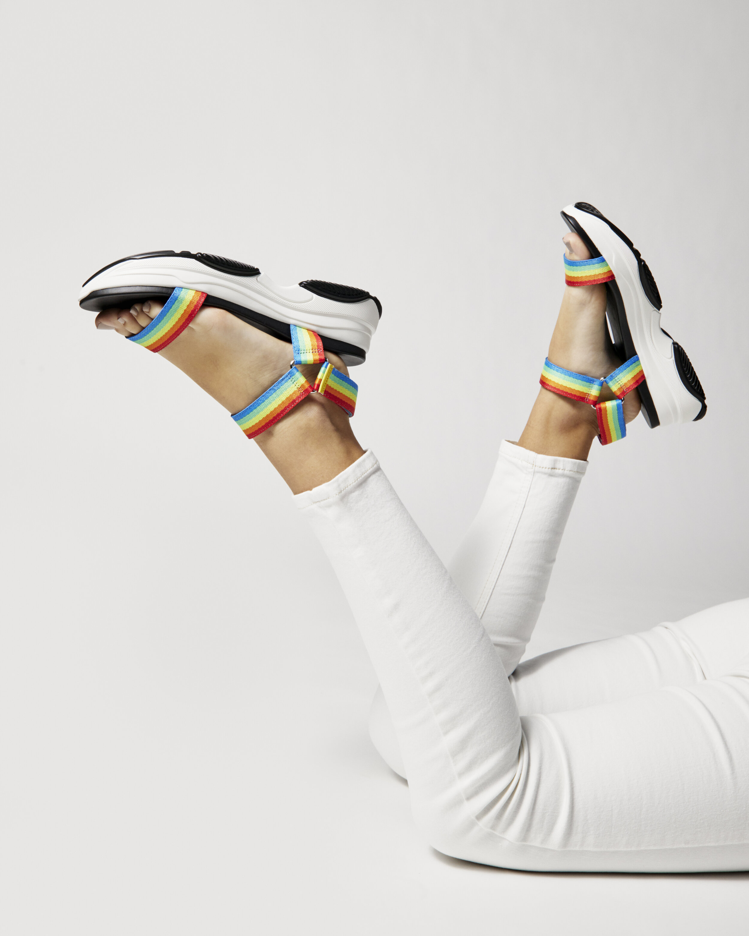 Person lying on their stomach with legs raised, wearing white shoes with rainbow straps, in white pants, against a plain light background.
