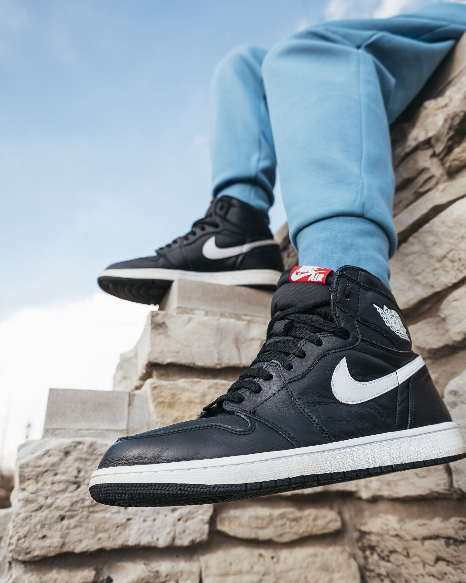 A person wearing black and white Nike Air Jordan sneakers and light blue pants, sitting on a stone wall outdoors with a clear sky in the background.