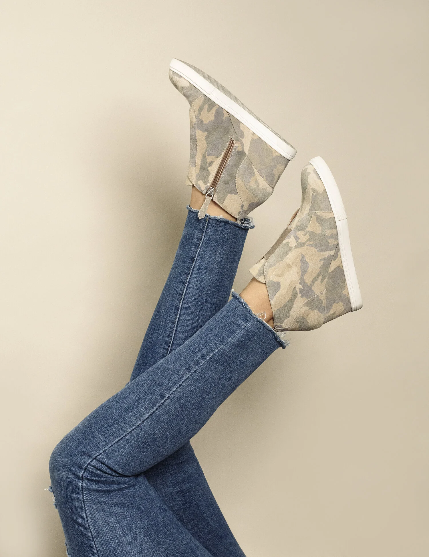 Person wearing blue jeans and camouflage boots lifting their legs against a plain wall.