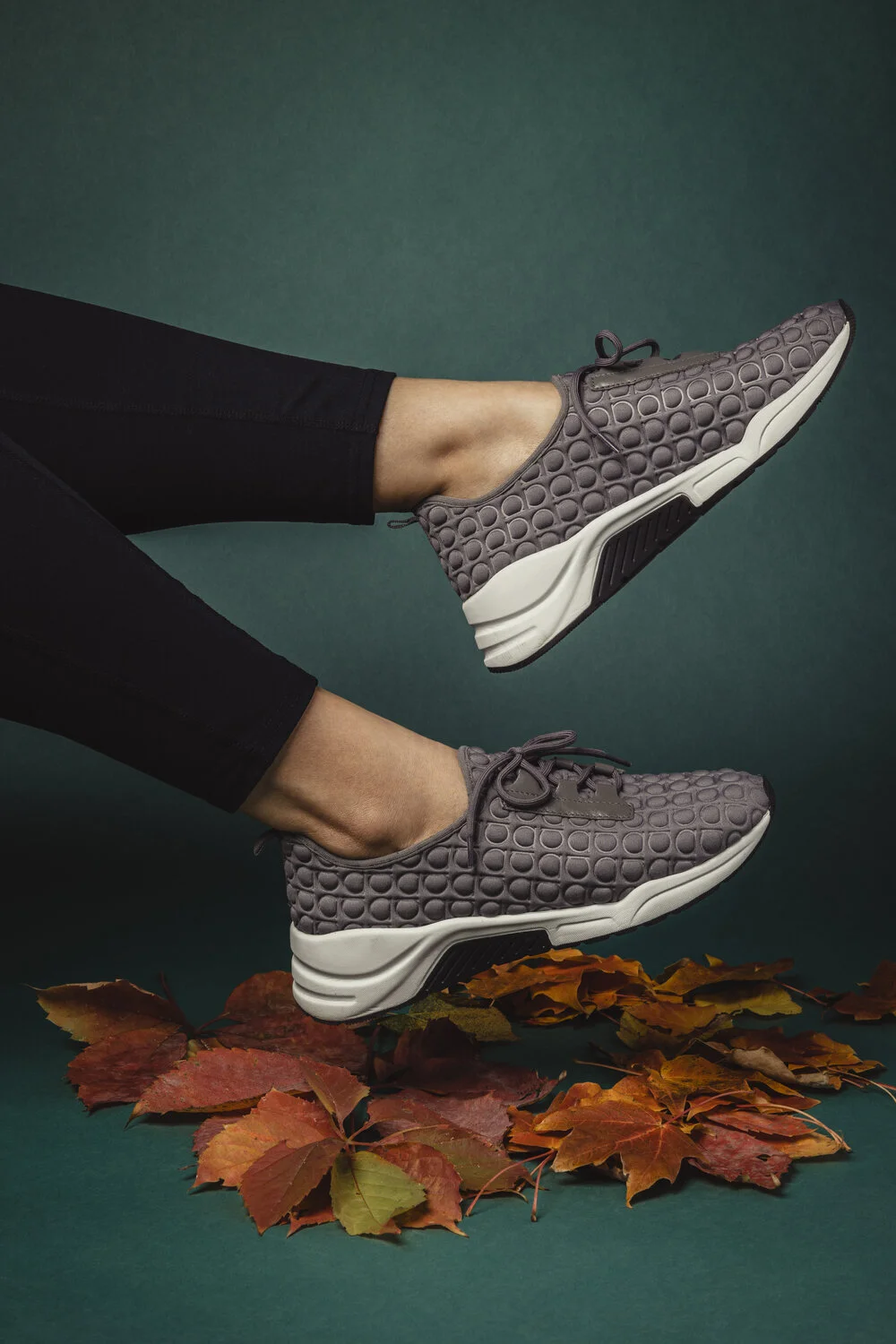 Person wearing gray textured sneakers and black pants, legs extended against a dark teal background, with autumn leaves on the ground.