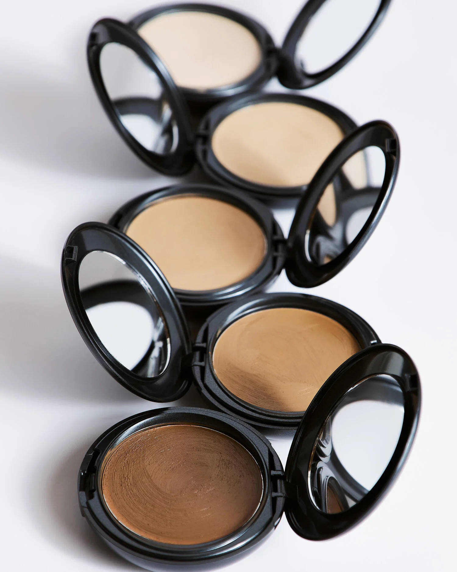 Four makeup compacts stacked in a diagonal line, each containing different shades of pressed powder foundation.