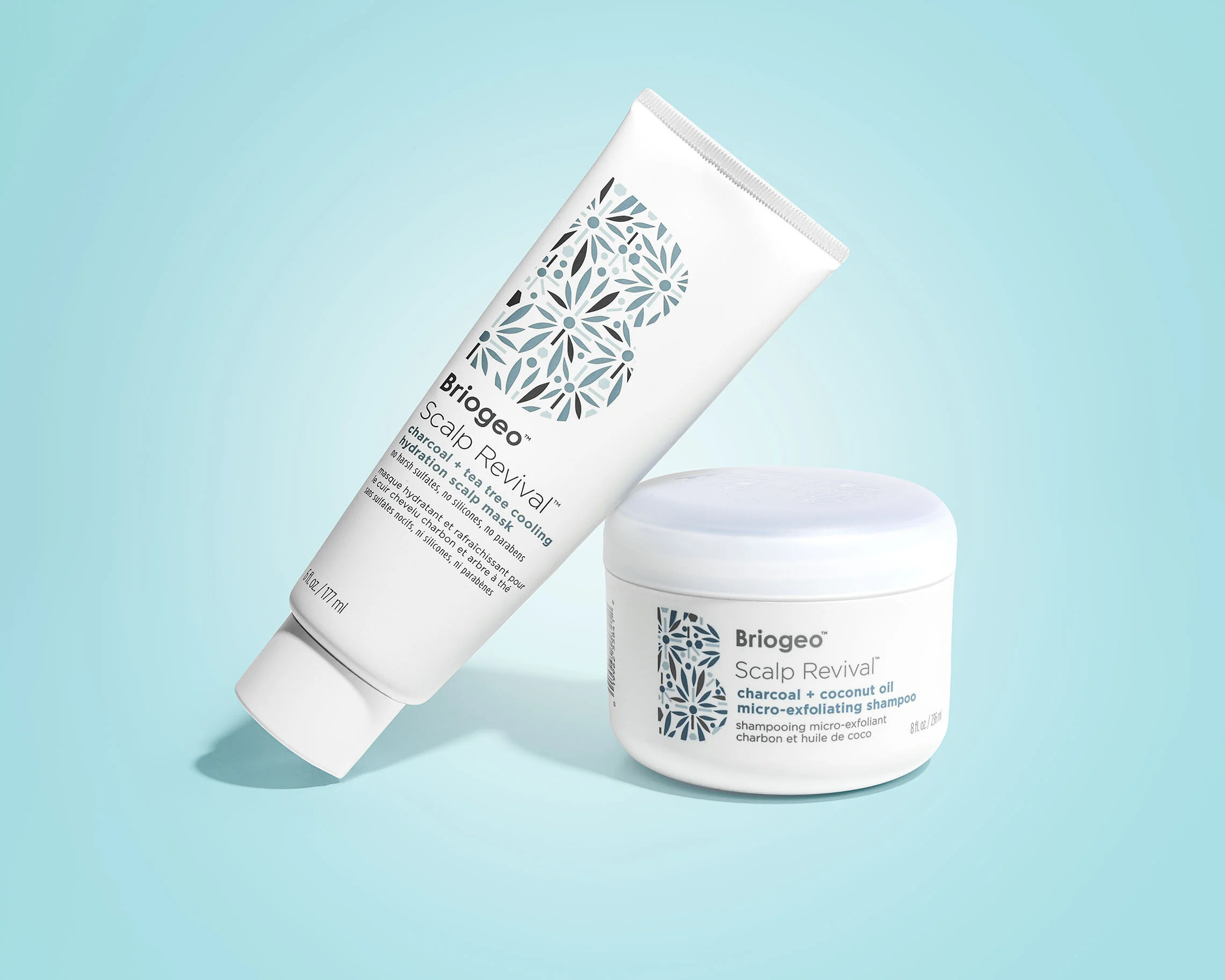 Two white hair care products from Briogeo on a light blue background: a tube of scalp mask and a jar of micro-exfoliating shampoo, both featuring blue and gray pattern designs.