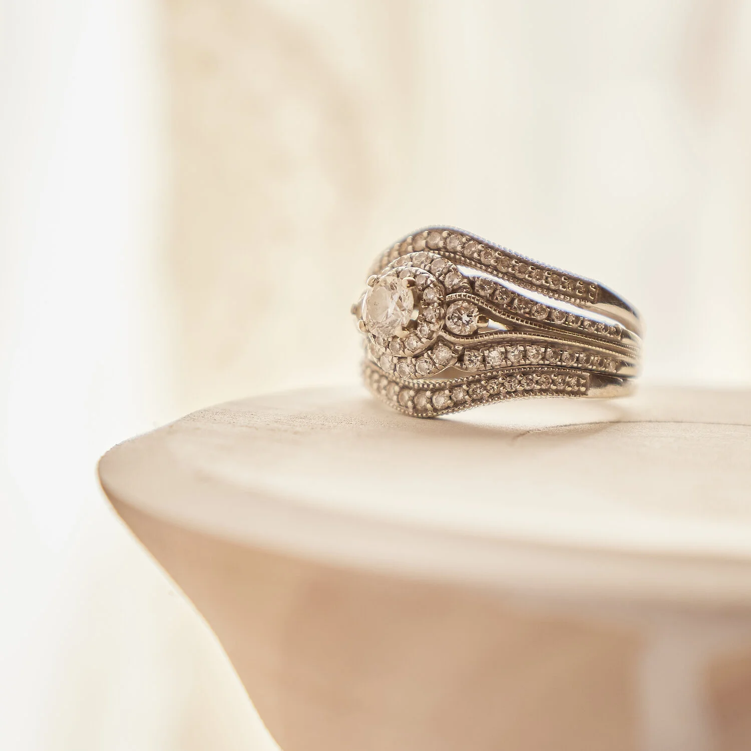 Close-up of a diamond ring with intricate band design, placed on a light-colored wooden surface.