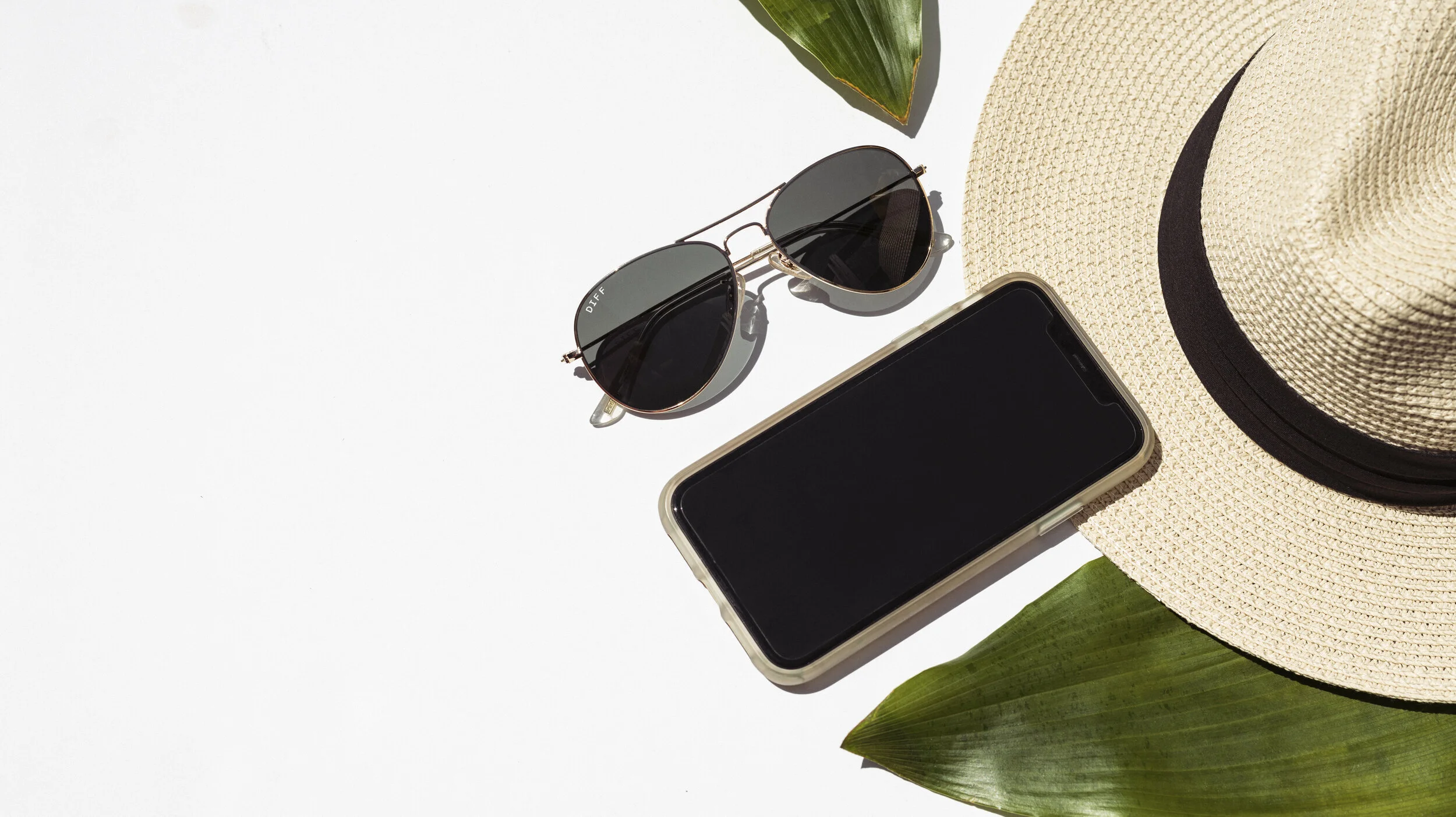 A flat lay of summer accessories including black sunglasses, a wide-brimmed straw hat, a smartphone with a black screen, and green leaves on a white background.