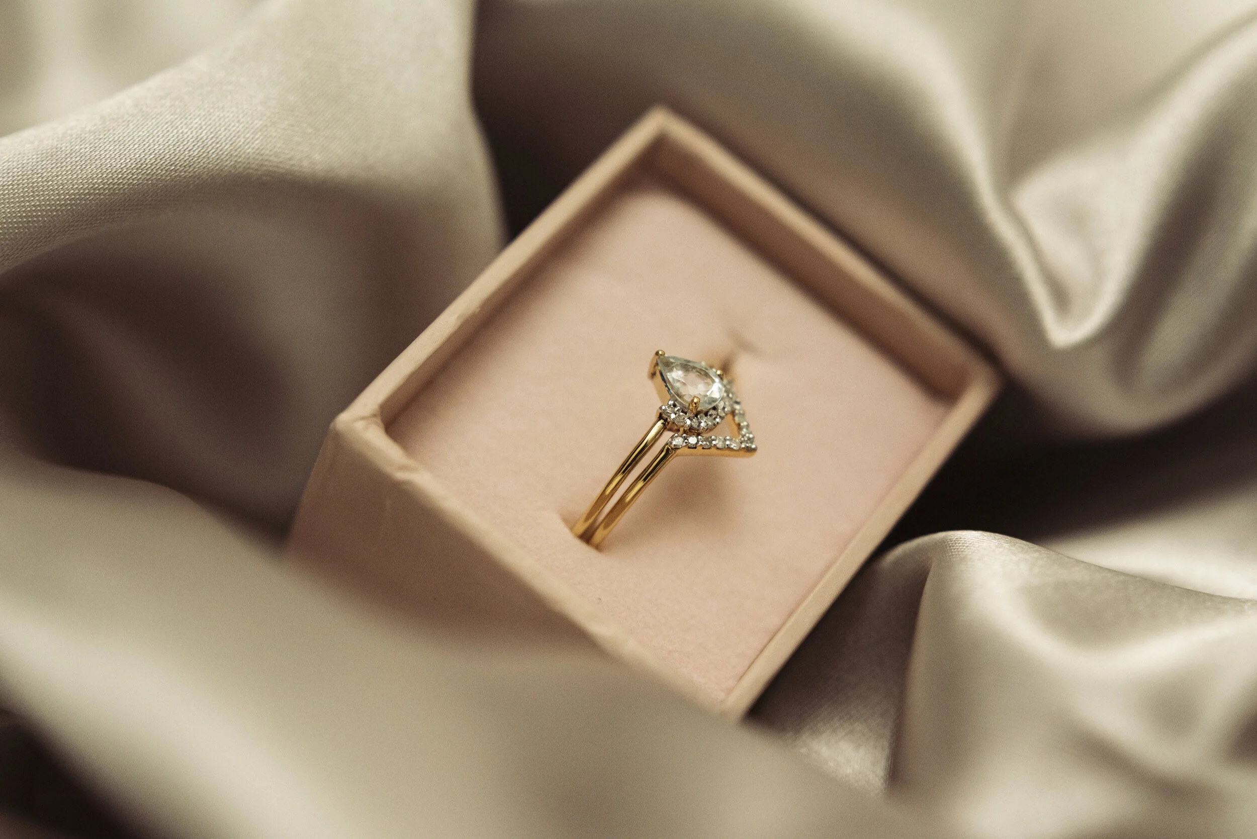 A gold ring with a pear-shaped diamond surrounded by smaller diamonds in a pink jewelry box, surrounded by satin fabric.
