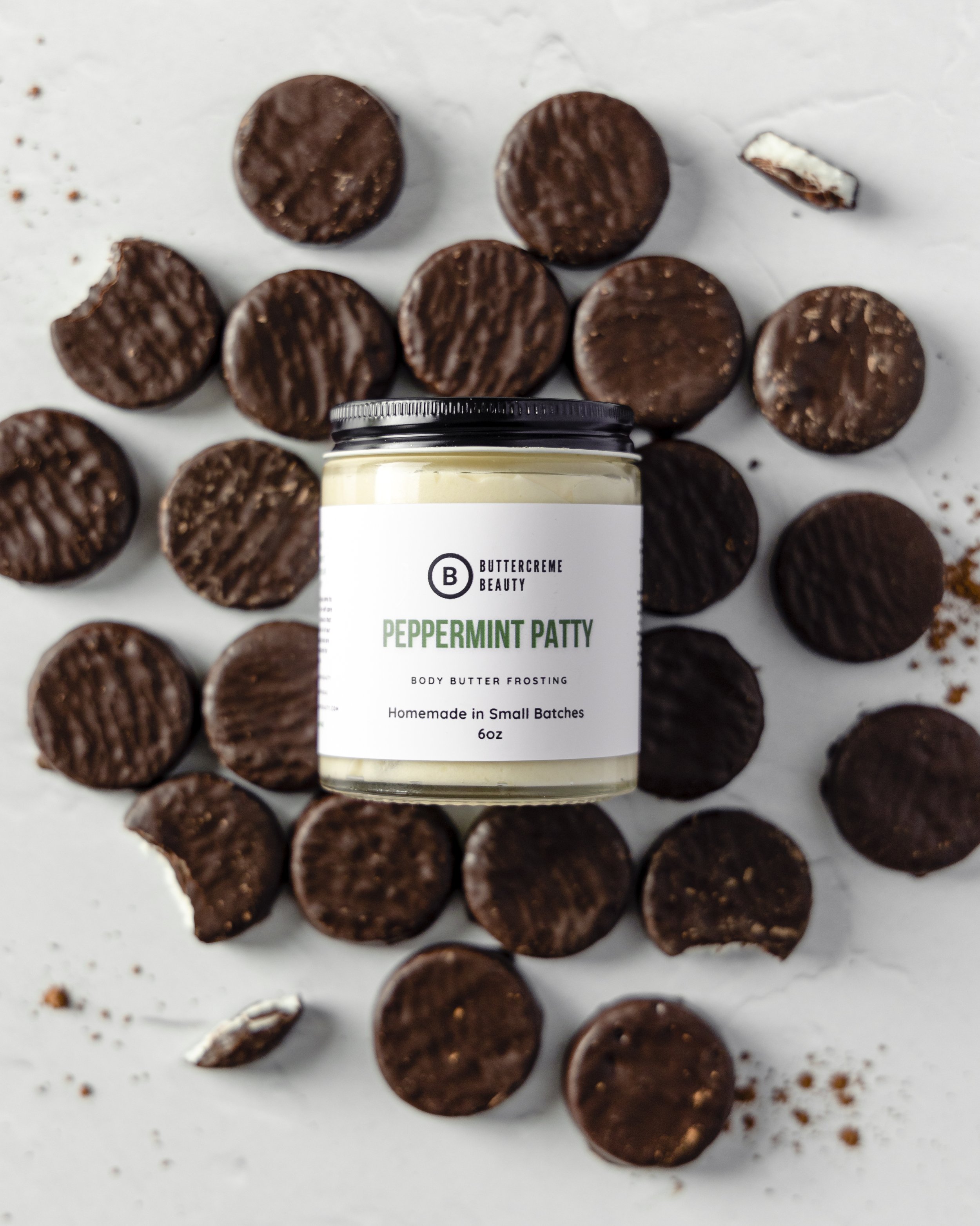 A jar of Buttercreme Beauty peppermint Patty body butter frosting surrounded by scattered peppermint patties on a white surface.
