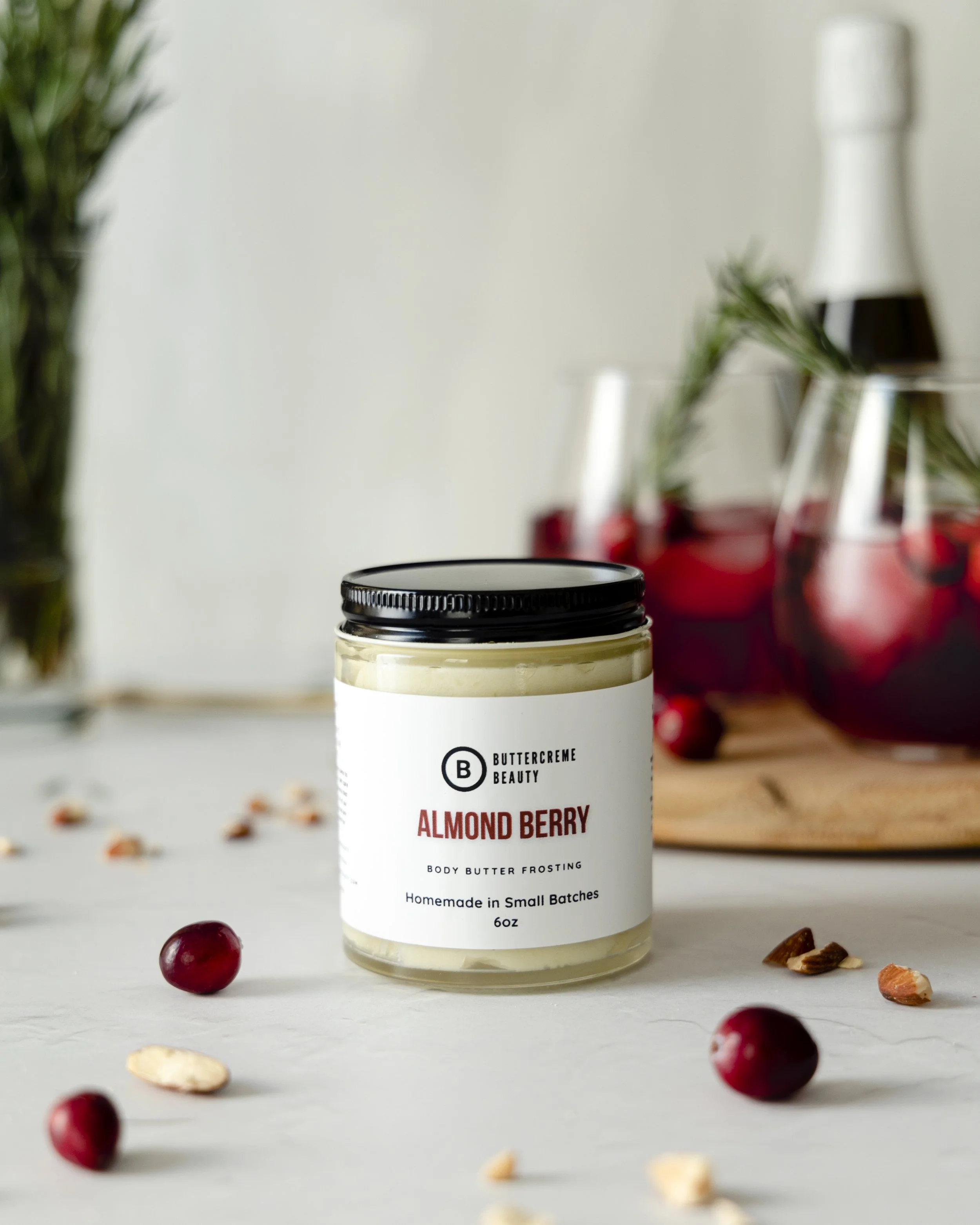 A jar of almond berry body butter frosting on a white surface, surrounded by cranberries and crushed nuts, with a blurred background of two glasses with red drink, fresh cranberries, and a white bottle.