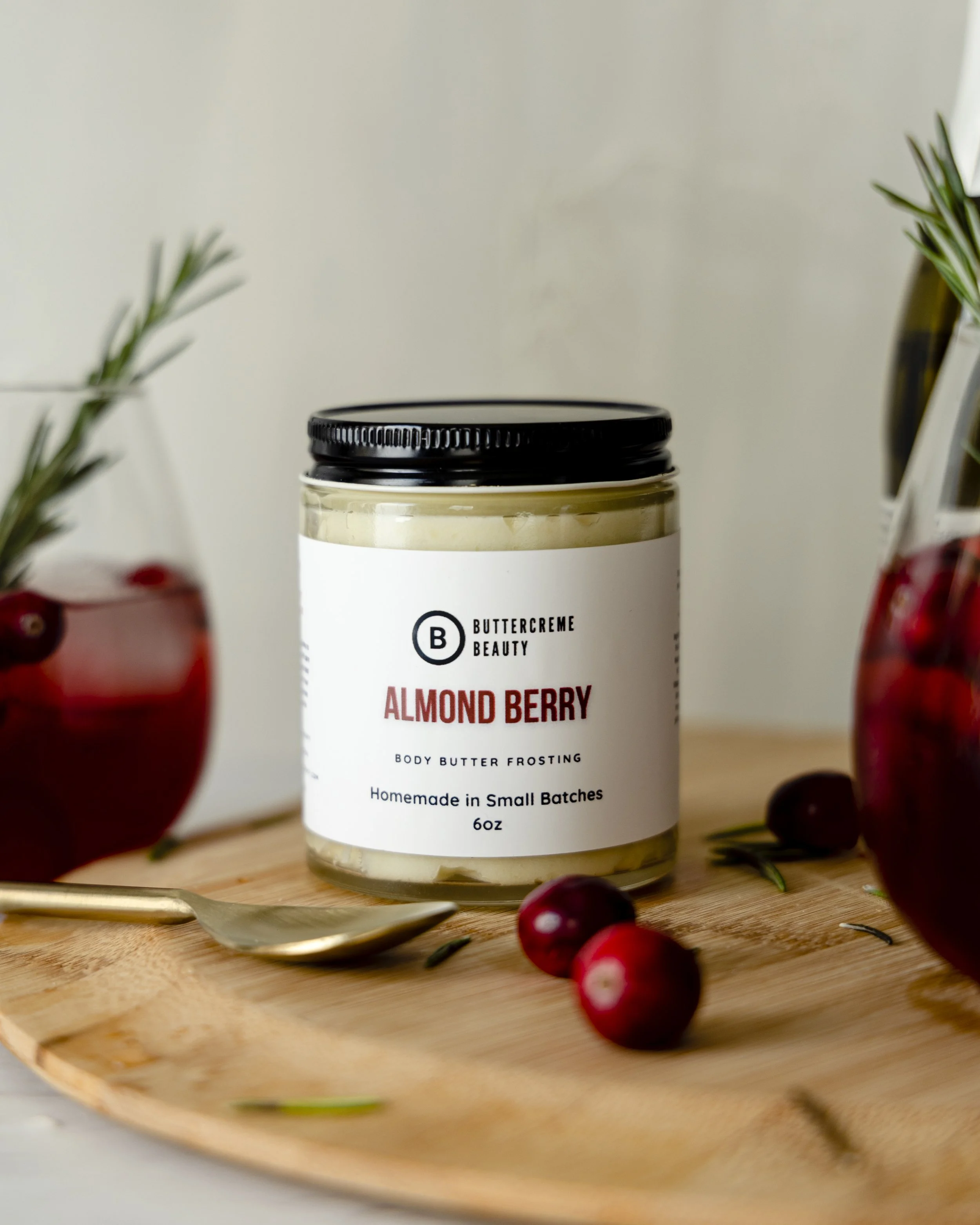 A jar of Almond Berry body butter frosting from Buttercrème Beauty on a wooden tray, accompanying red berries and greenery in vases.