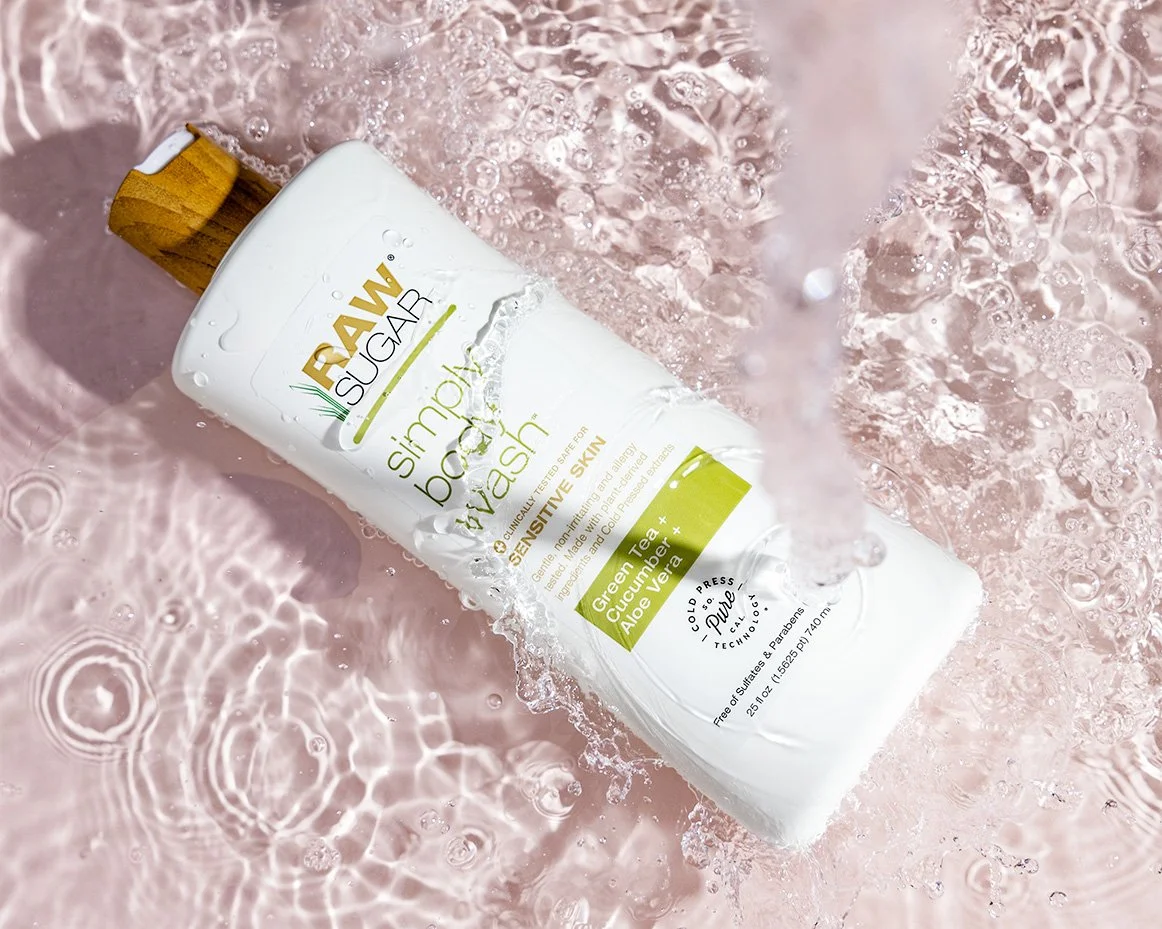 A bottle of RAW Sugar simple body wash with green tea, cucumber, and aloe vera sitting on a pink surface with water splashing around it.