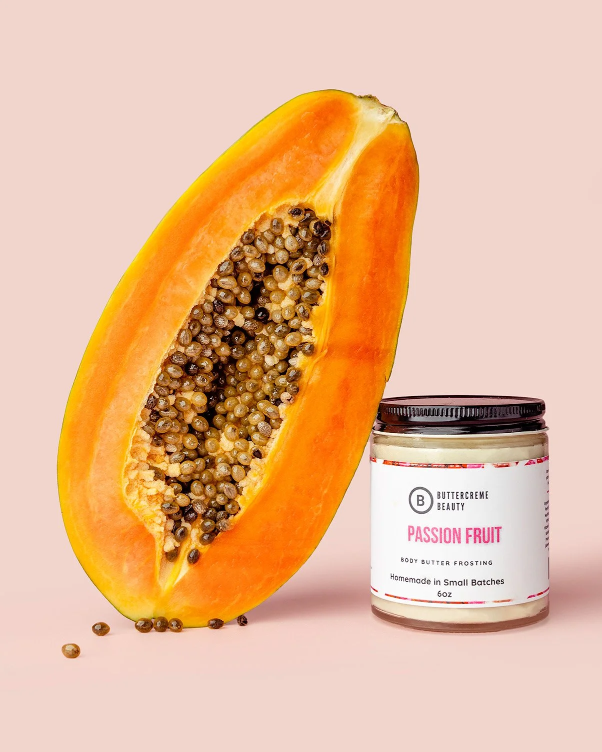 Half of a ripe papaya with orange flesh and black seeds next to a jar of passion fruit body butter frosting with a white label and black lid on a light pink background.