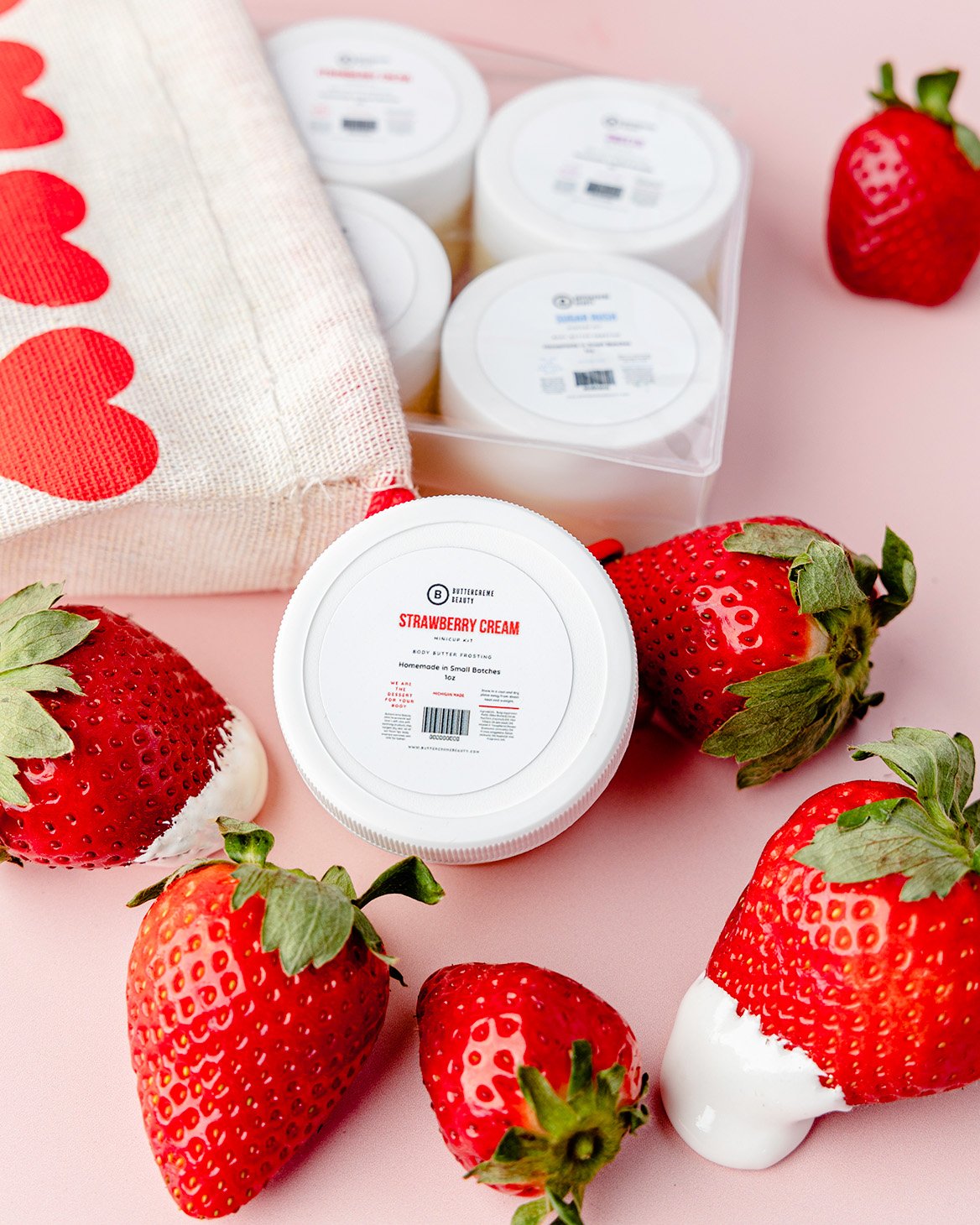 Fresh strawberries with white cream or frosting, strawberry flavored cream in containers, a bag with red hearts printed on it, and a pink background.