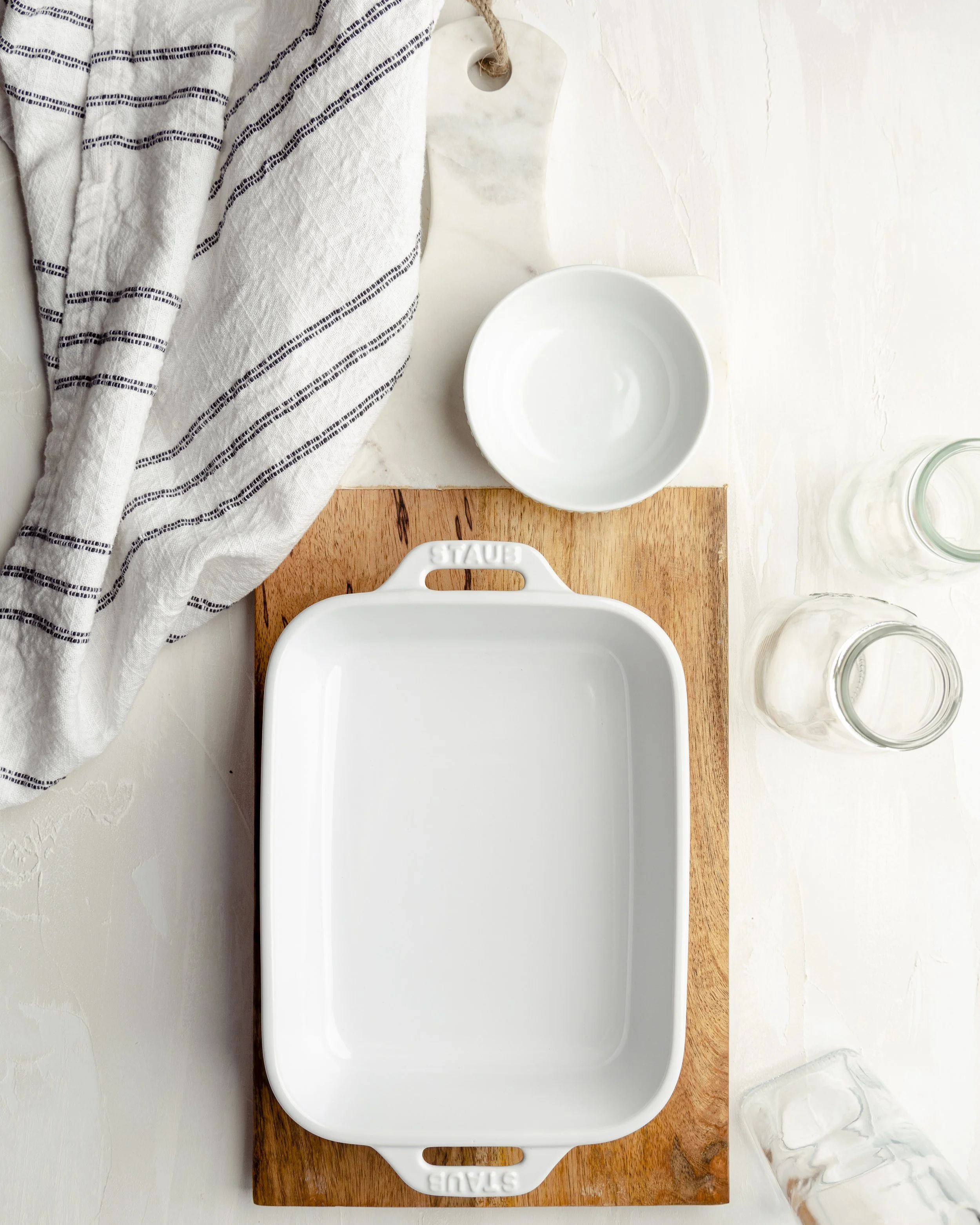 Empty white baking dish on a wooden cutting board, with a white bowl and two glass jars nearby, a dish towel on the left, and a glass of water at the bottom right.