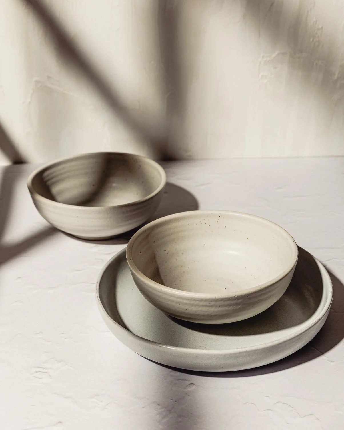Three white ceramic bowls, one placed on a matching ceramic plate, with shadows cast on a white textured surface and background.