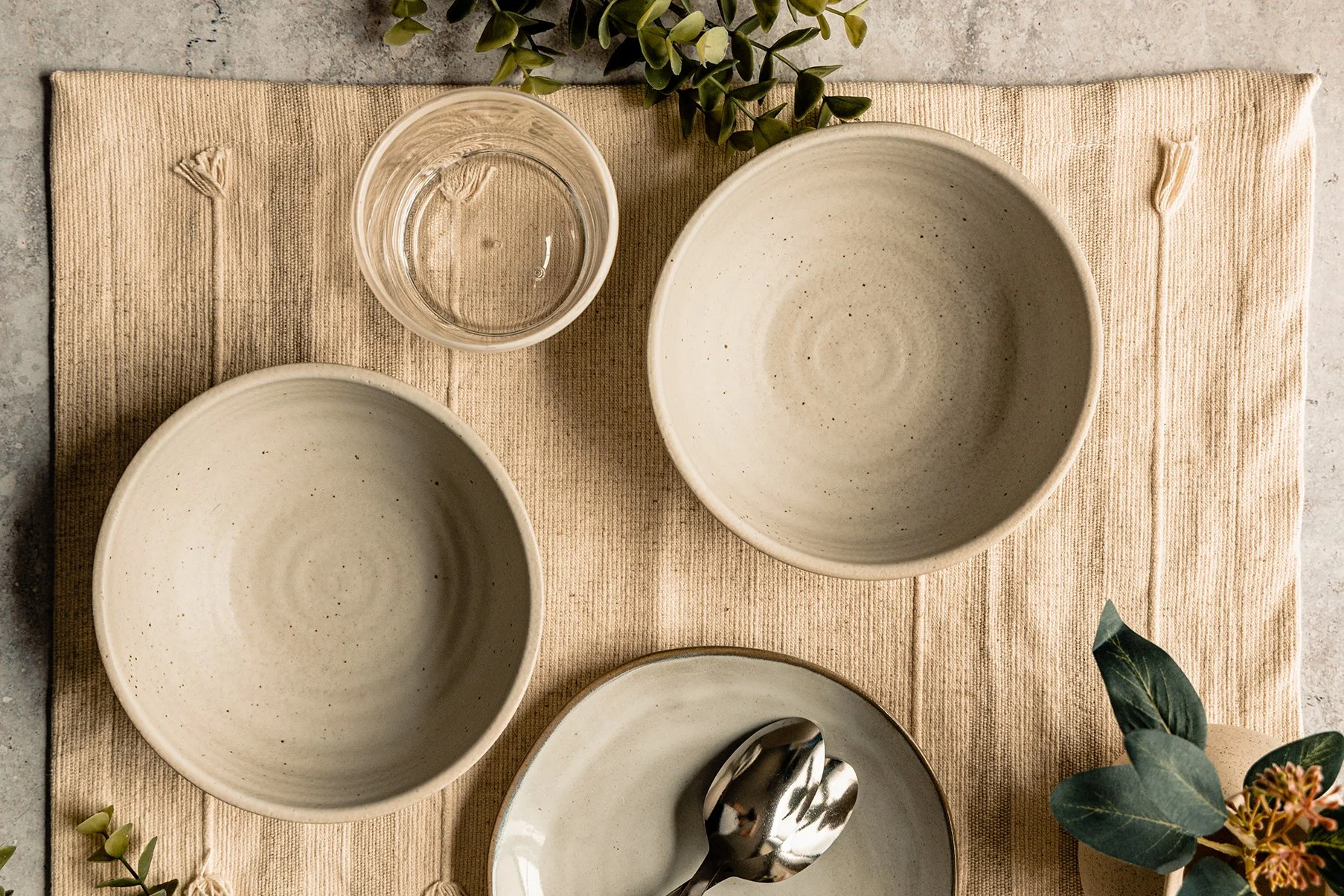 A top-down view of a dining table setting with beige bowls, a glass of water, a plate with two spoons, and green foliage on a beige textured cloth and gray surface.
